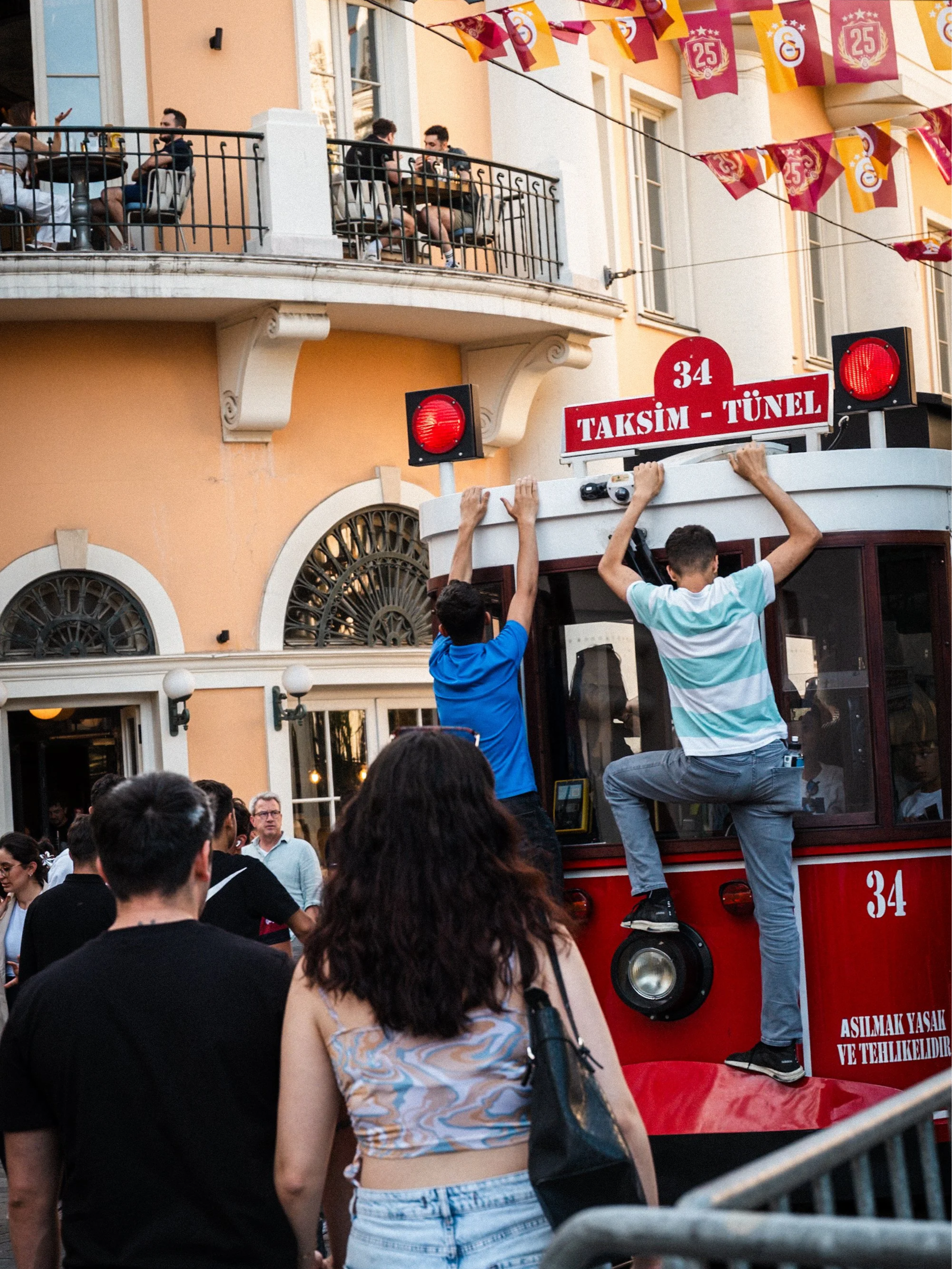 Teenagers hanging onto the back of the historic red Taksim–Tünel tram on İstiklal Avenue in Istanbul, surrounded by crowds and flags — dynamic Istanbul street photography capturing the energy of the famous pedestrian street.
