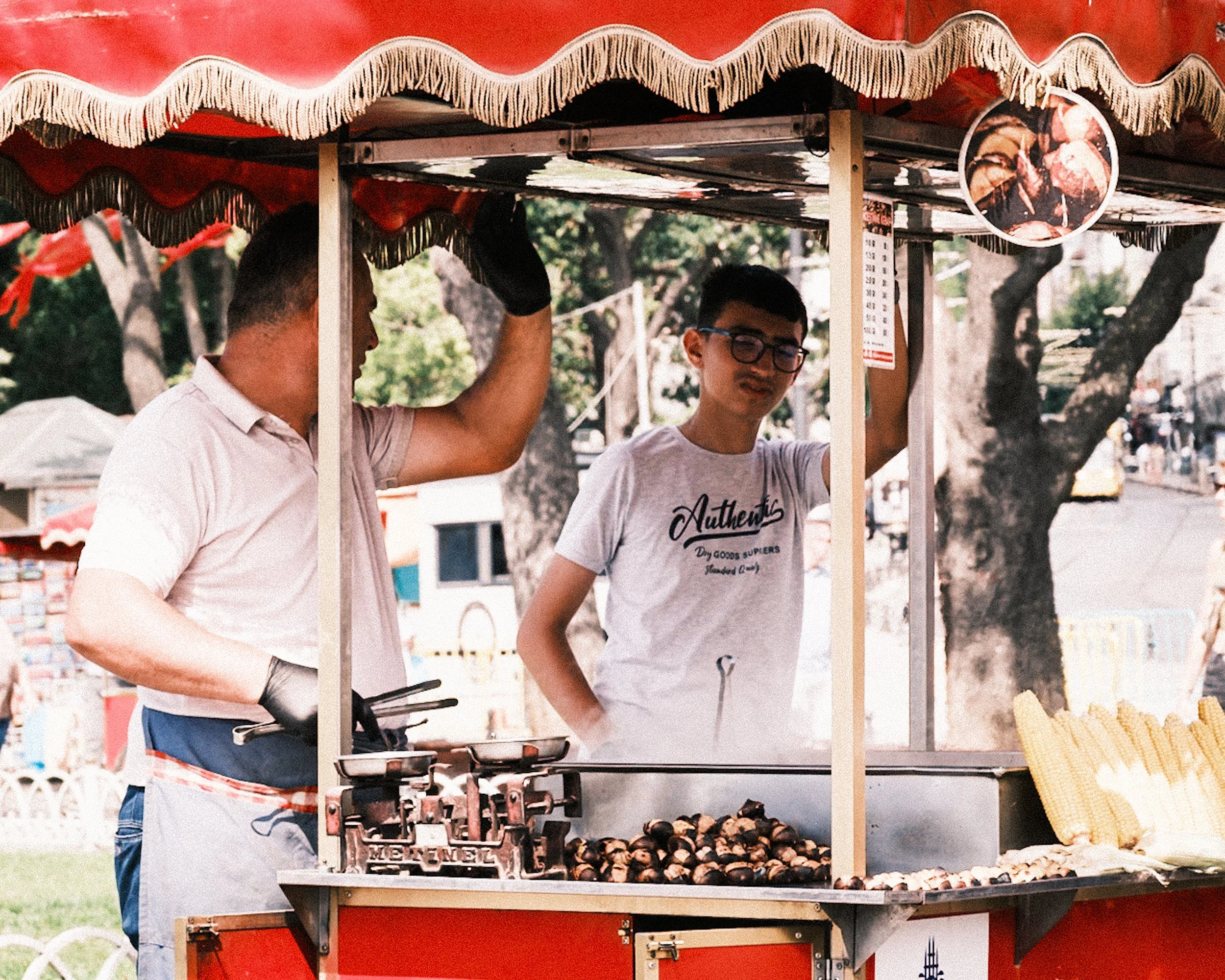 A traditional chestnut vendor works behind his red street cart in Istanbul while a young customer watches the roasting process. The warm smoke, old-fashioned weighing scale, and vibrant colors of the cart highlight the timeless charm of Turkish stree