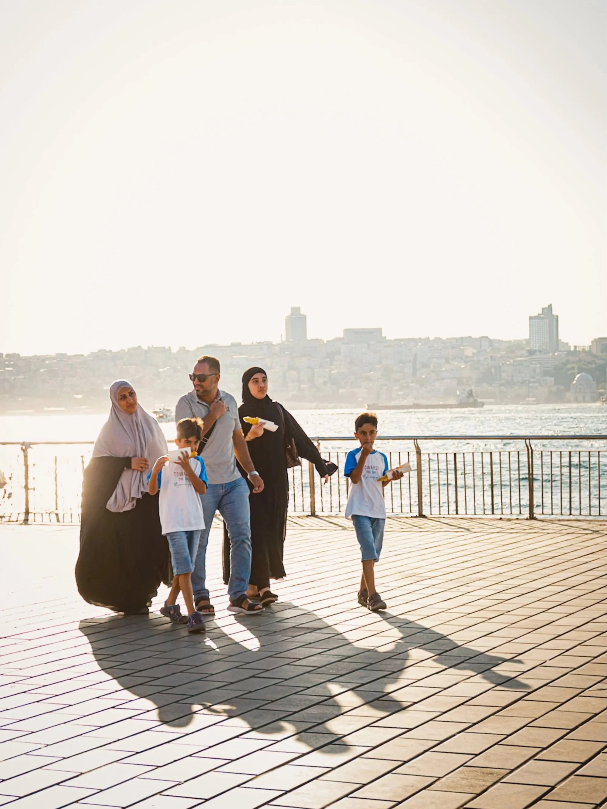 Family walking along the waterfront near the Bosphorus in Istanbul during golden hour, with long shadows stretching across the pavement and the city skyline glowing in the background — warm, cinematic Istanbul street photography.