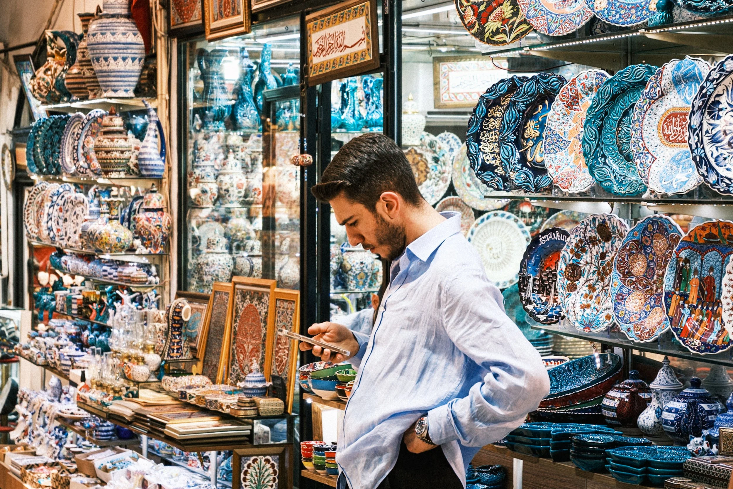 Young man looking at his phone inside a traditional Istanbul ceramics shop filled with colorful handmade pottery, tiles, and decorative plates — vibrant Istanbul street photography in the Grand Bazaar area.