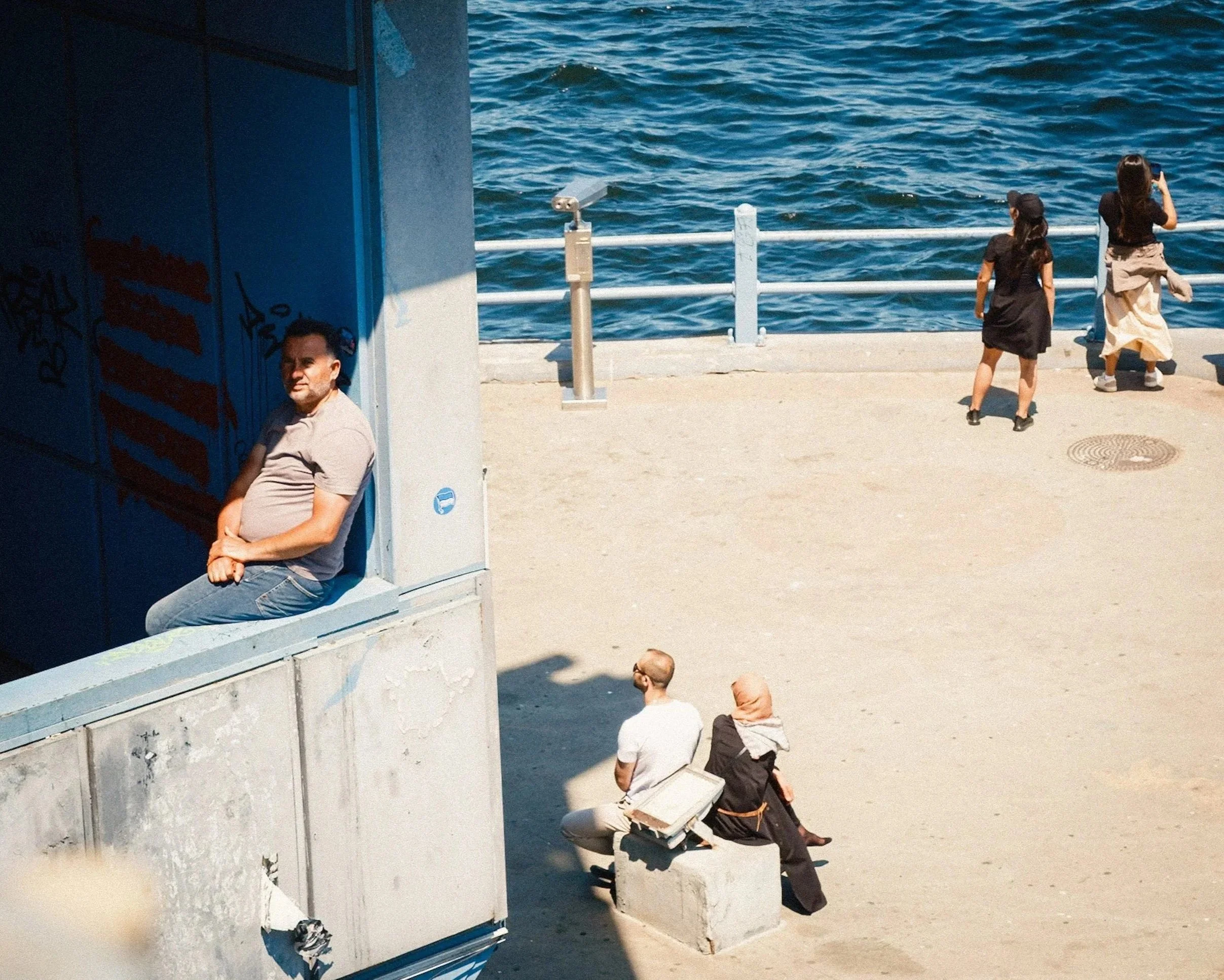 Man sitting in the shaded corner of an underpass near the waterfront while other people enjoy the sunny promenade by the Bosphorus in Istanbul — minimalist Istanbul street photography showing contrasts of light, shadow, and movement.