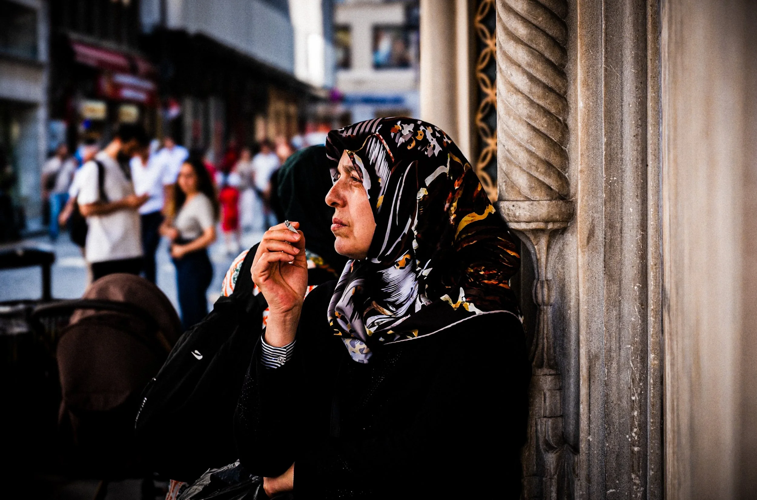 Woman wearing a patterned headscarf standing by a carved stone column and smoking while watching the crowded street in Istanbul — expressive Istanbul street photography capturing emotion and urban life.