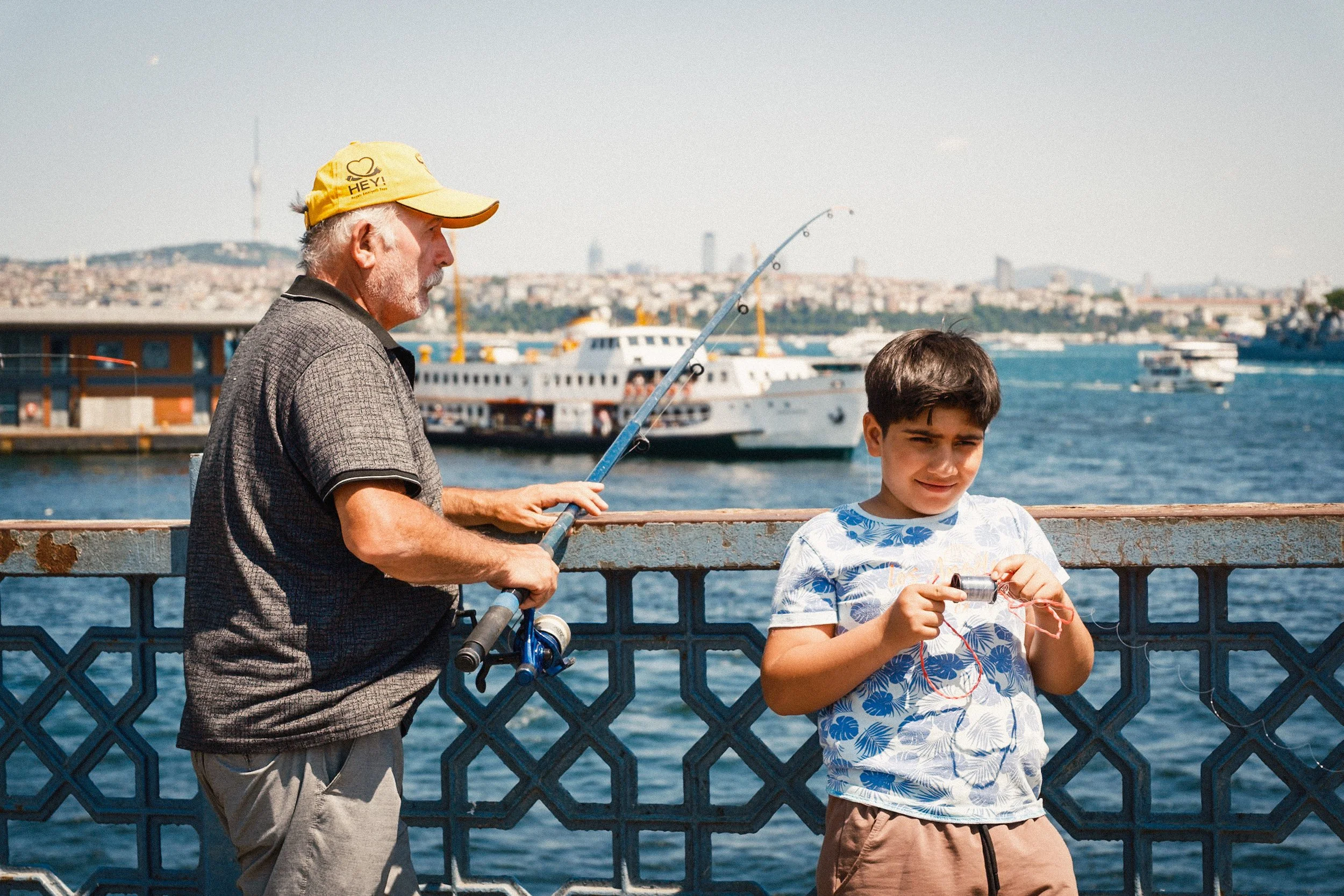 Older man fishing from the Galata Bridge while a young boy stands beside him holding fishing line, with ferries and the Istanbul skyline in the background — warm Istanbul street photography showing family and local fishing traditions.