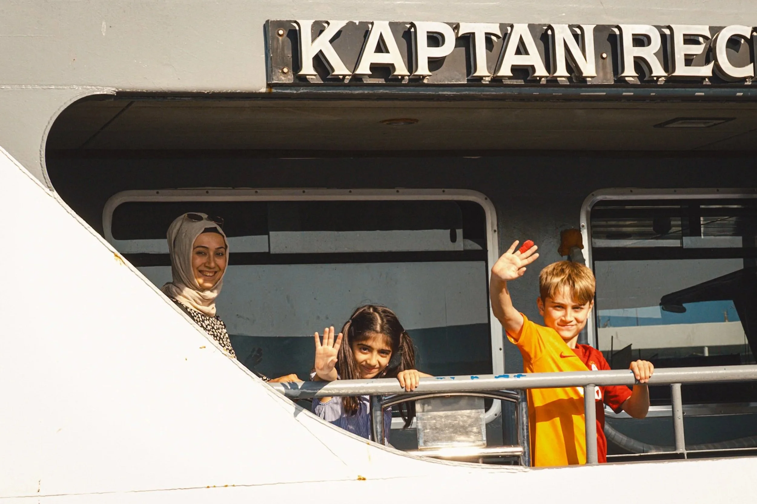 Children waving cheerfully from a ferry alongside a smiling woman as the boat departs from the dock in Istanbul — joyful Istanbul street photography showing travel moments and life on the Bosphorus.