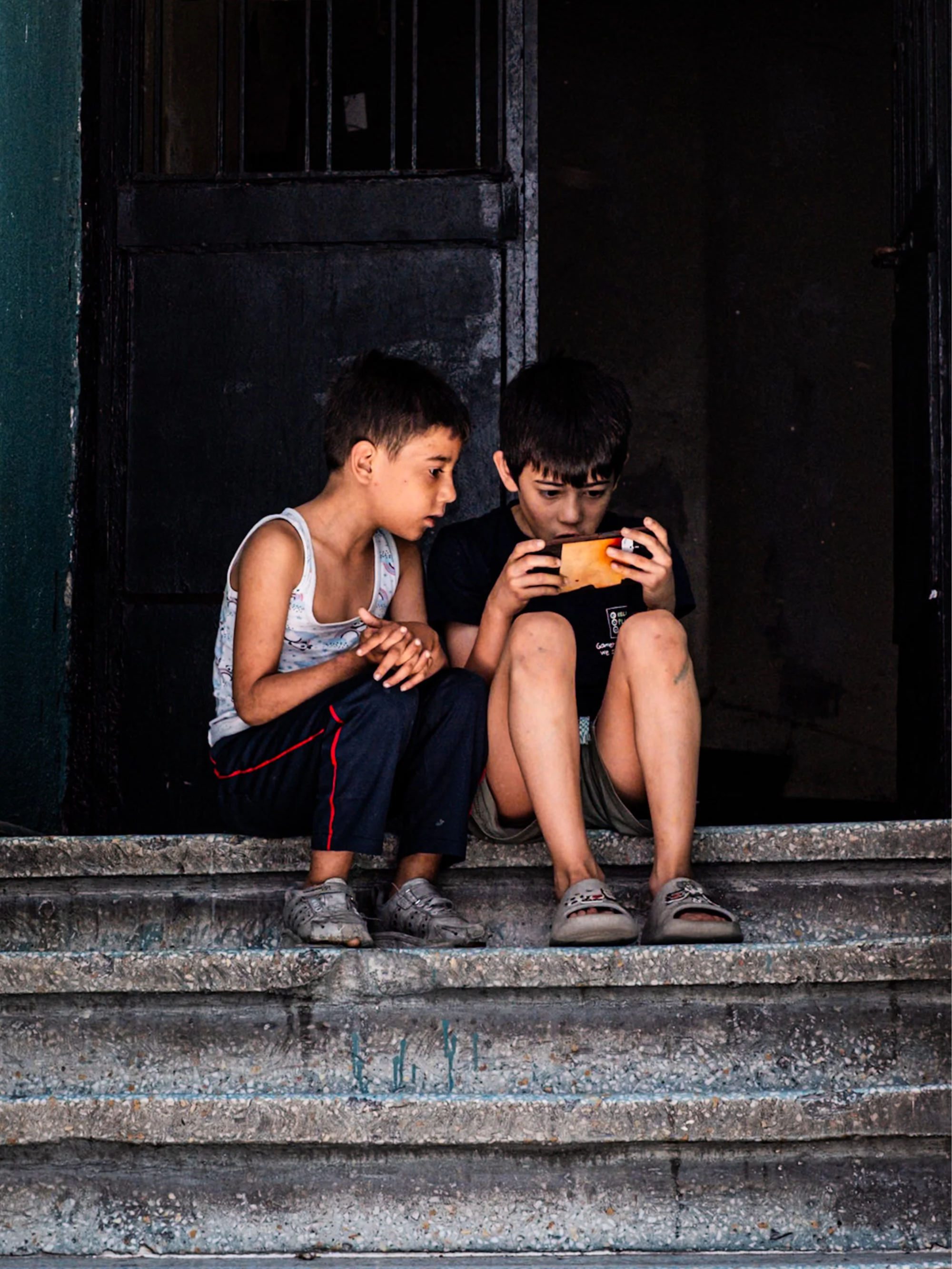Two young boys sitting on worn concrete steps, one focused on a mobile phone while the other watches closely — intimate Istanbul street photography showing everyday childhood moments in a neighborhood alley.