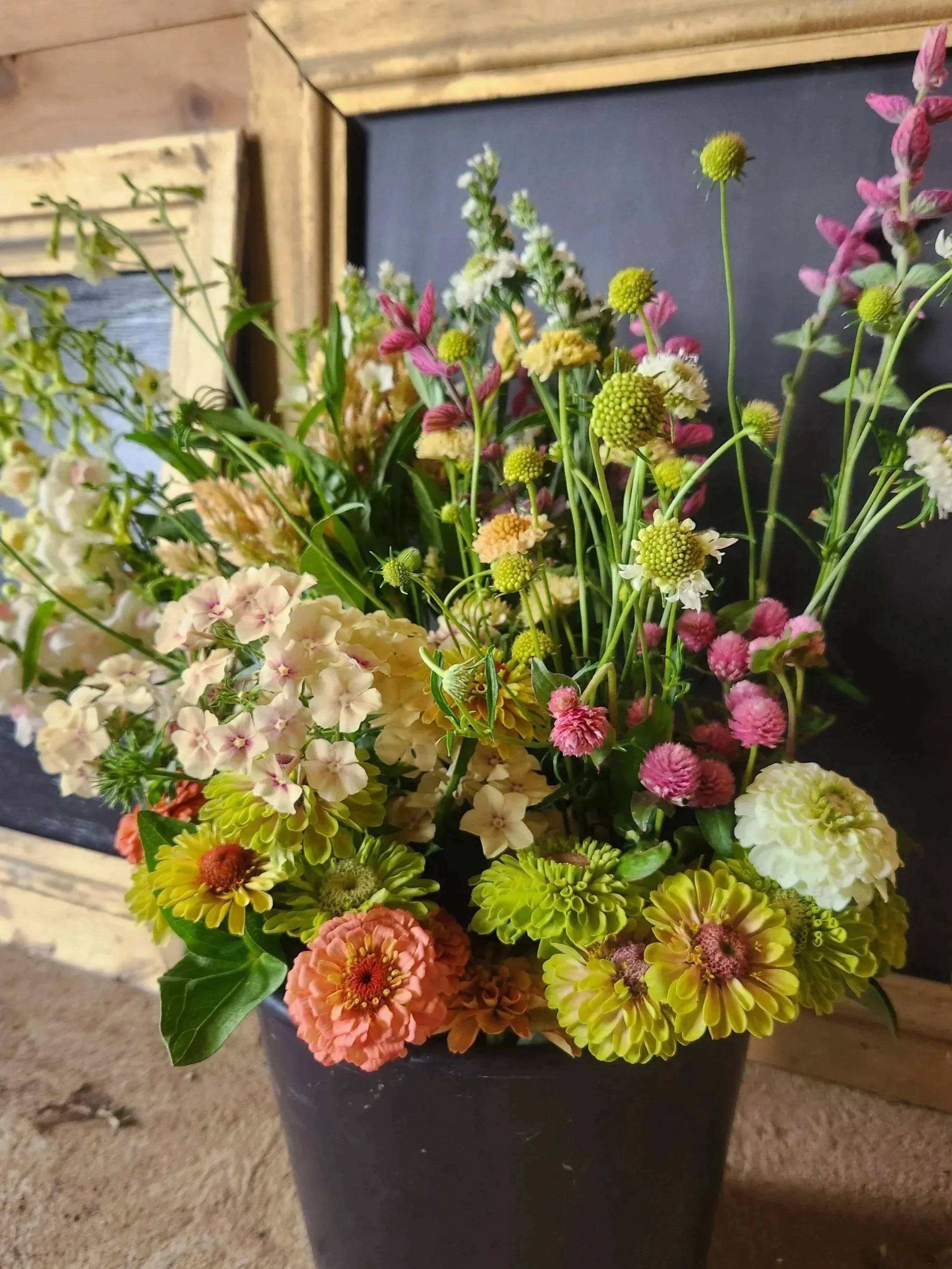 A colorful bouquet of various flowers in a black pot placed on a wooden surface, with a wooden frame and chalkboard in the background.