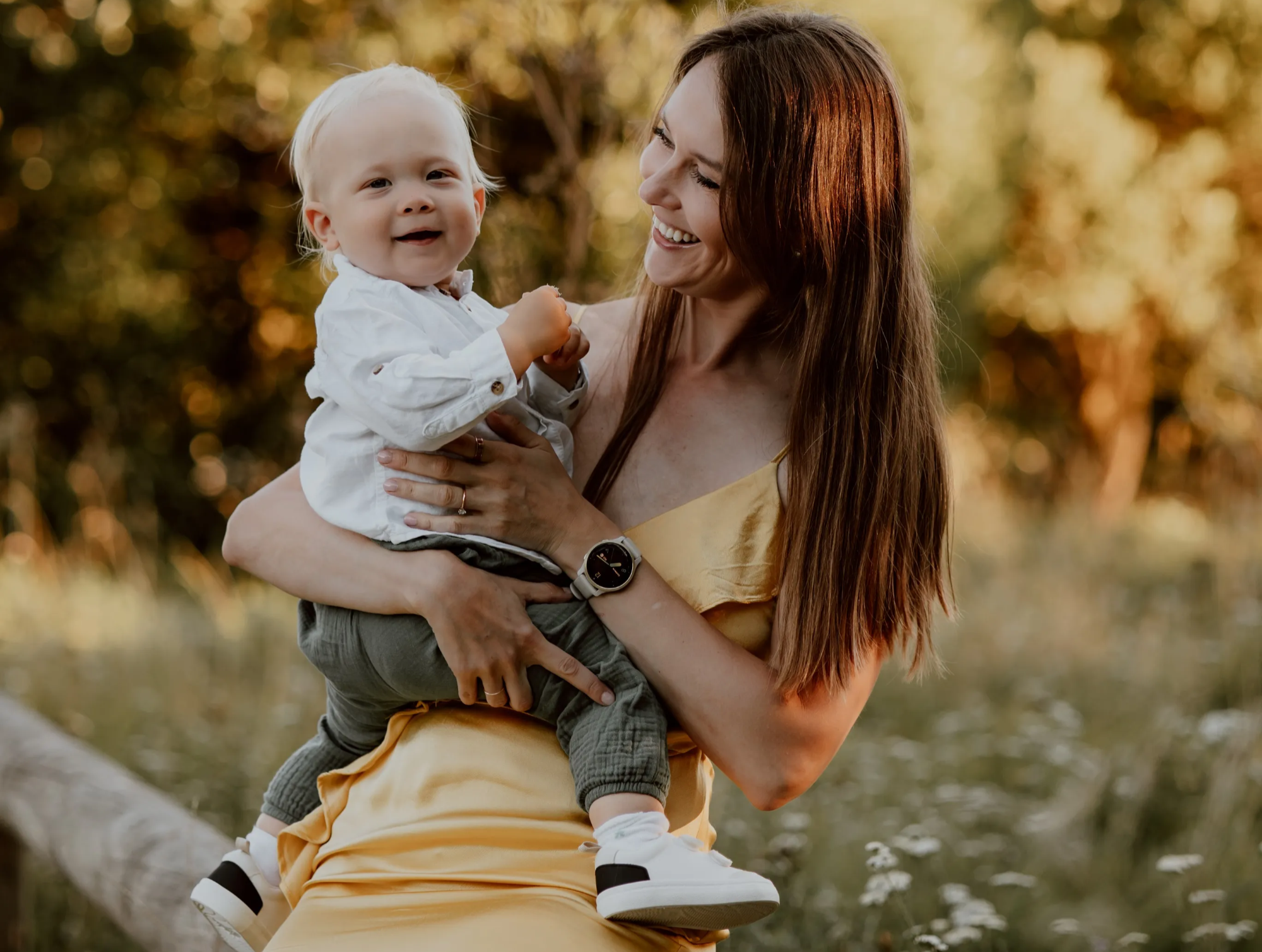 A woman with long reddish hair holding a smiling toddler boy with blonde hair outdoors during sunset, with trees and water in the background.
