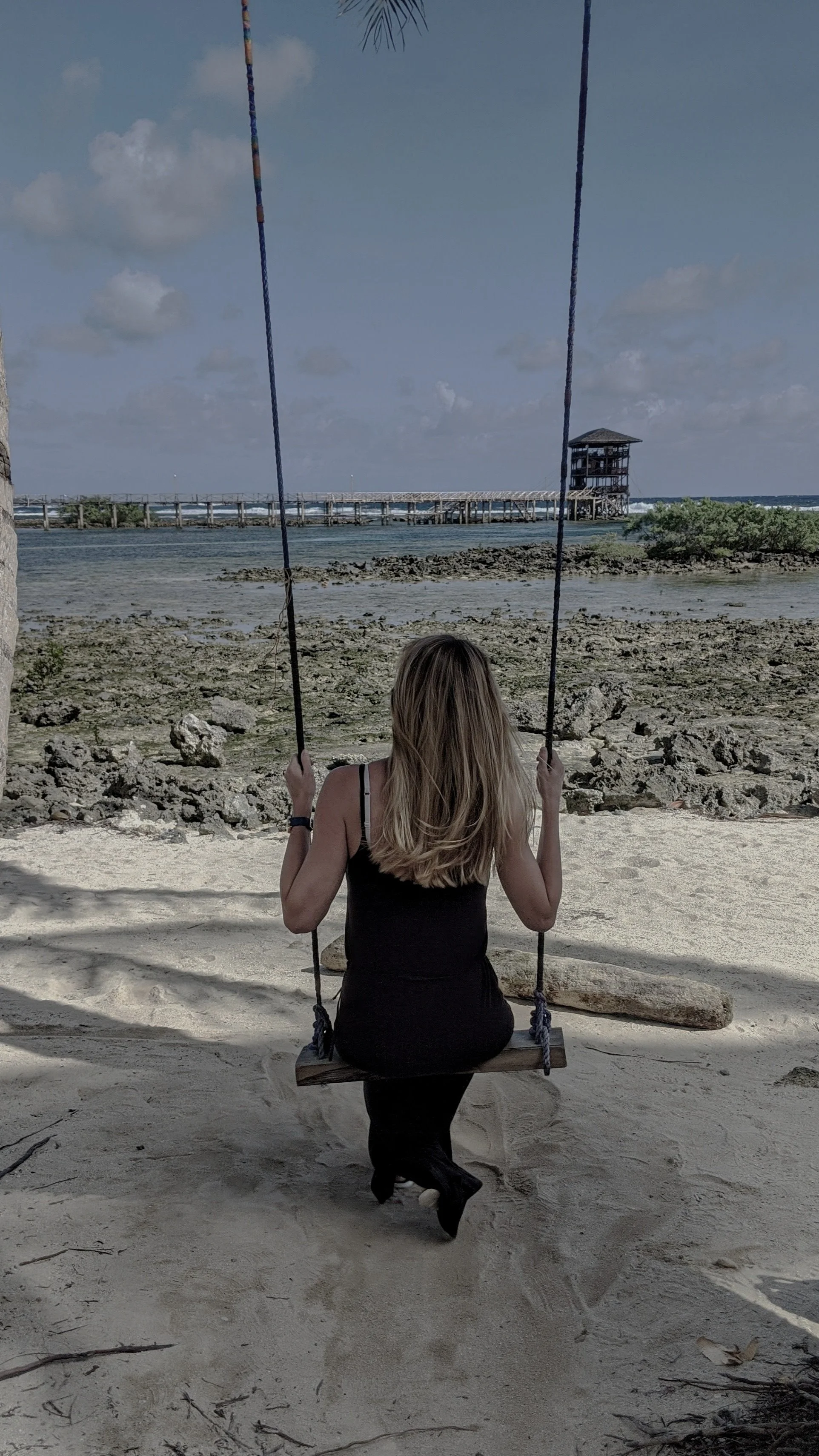 A woman with blonde hair sitting alone on a wooden swing at the beach, facing the water and a pier in the distance under a partly cloudy sky.
