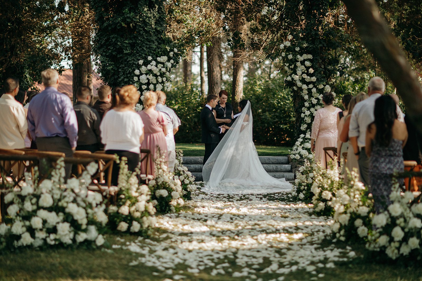 Outdoor wedding ceremony with bride and groom beneath a flower-covered arch and guests seated along a petal-lined aisle