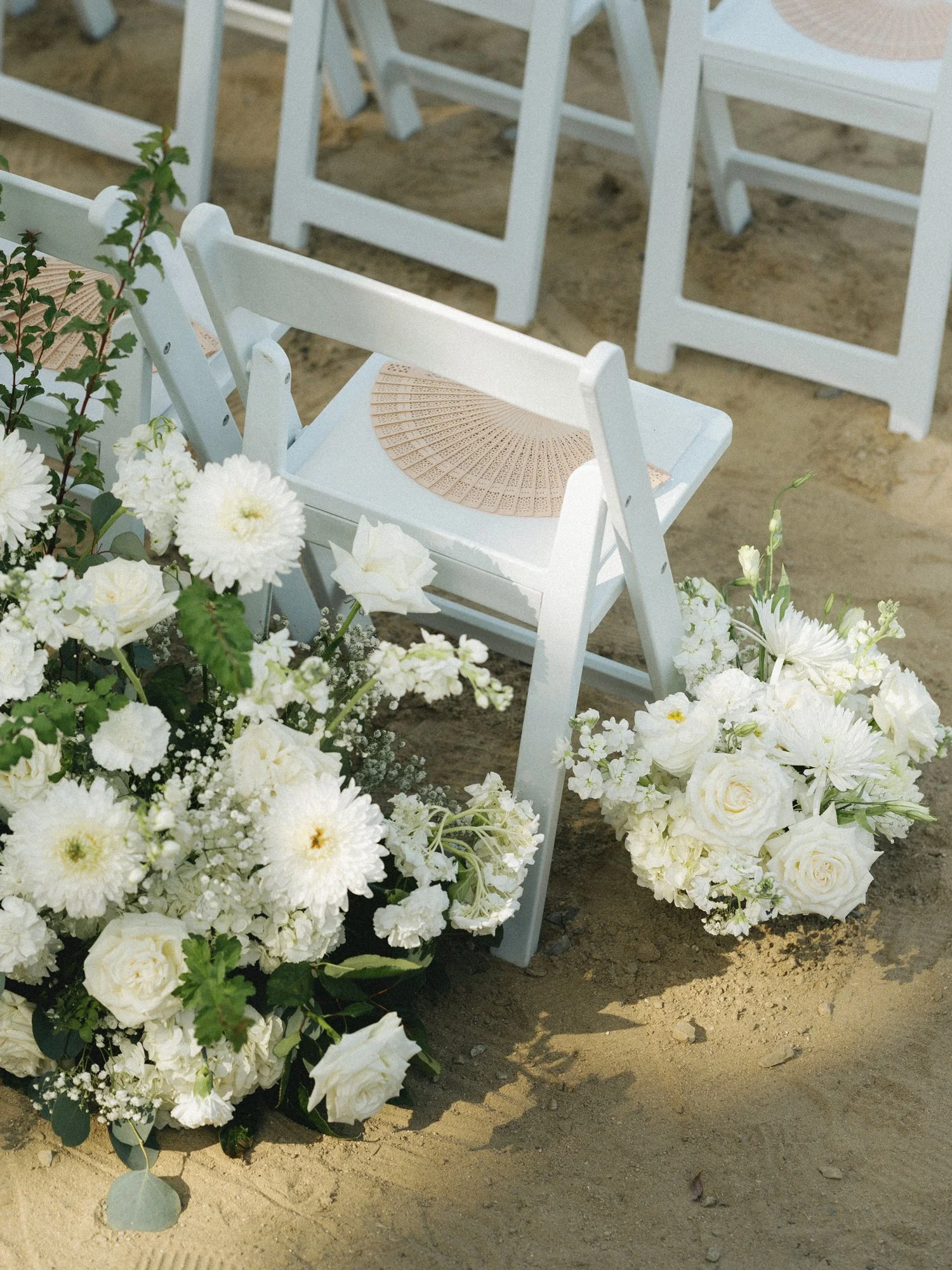 White folding chairs arranged on sandy ground with a woven fan and white floral wedding arrangements.