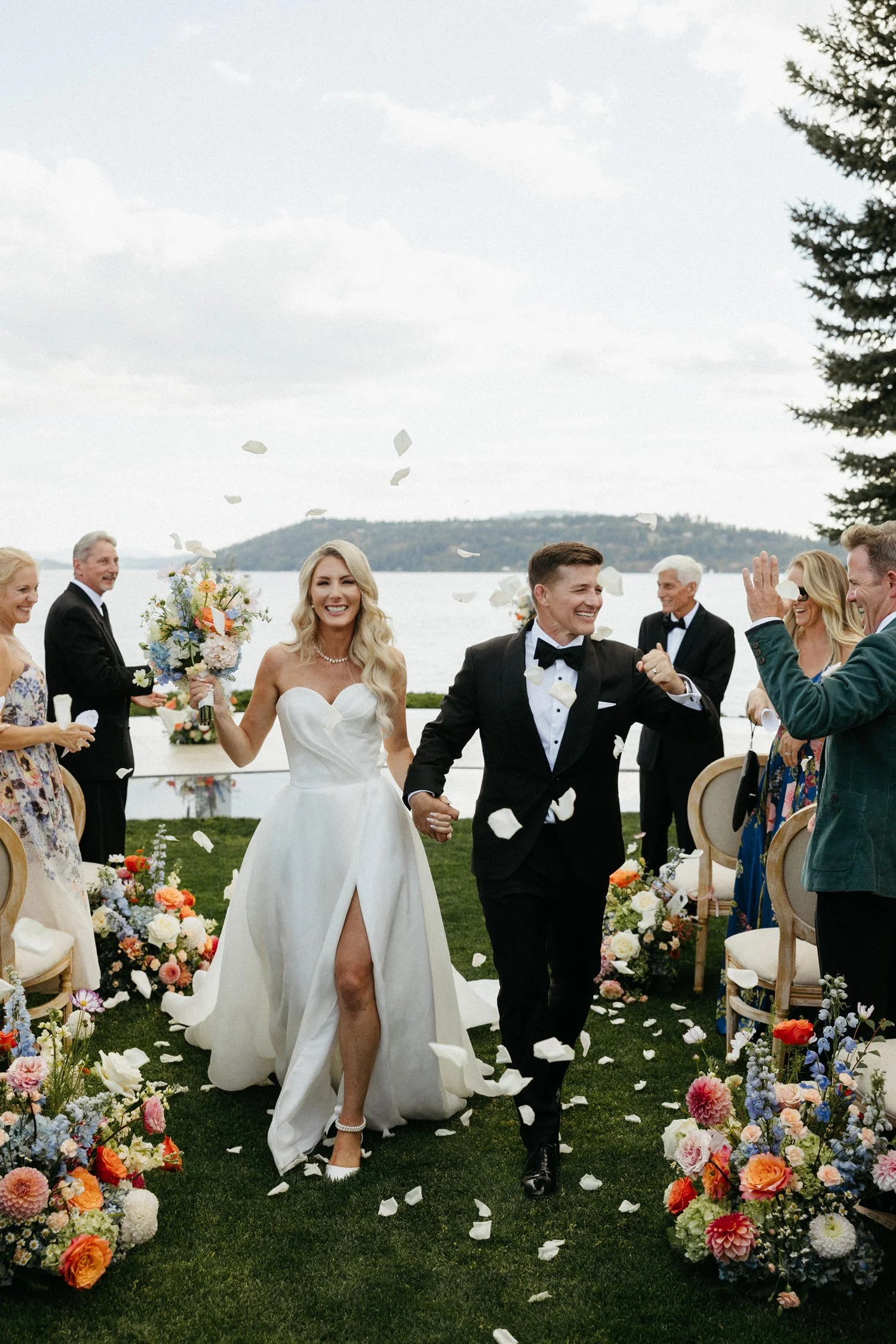 Bride and groom celebrating their outdoor lakeside wedding as guests toss flower petals among colorful floral arrangements.