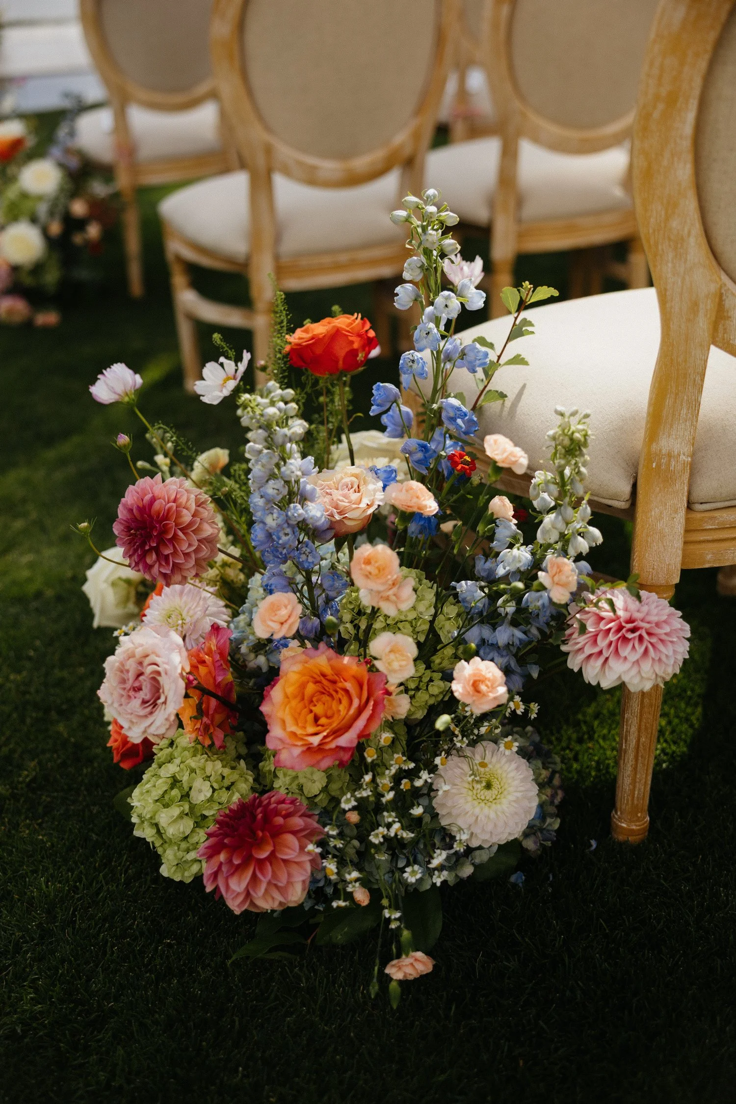 Colorful floral arrangement with roses, dahlias, delphiniums, hydrangeas, and daisies beside rattan chairs at an outdoor event