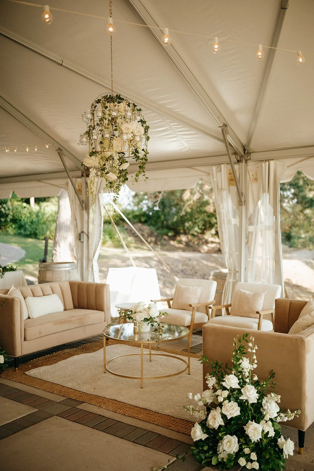 Elegant outdoor wedding tent lounge with cream sofas, armchairs, floral arrangements, a glass coffee table, and a hanging floral chandelier.