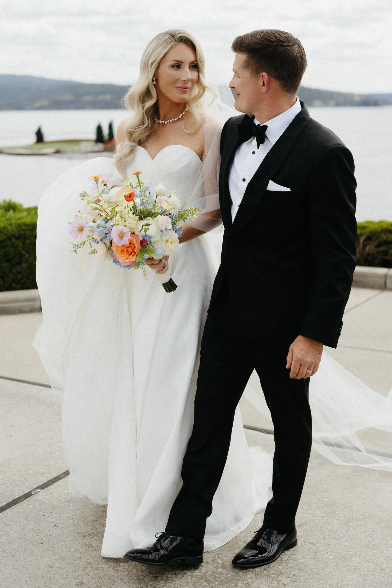 Bride in a white wedding gown holding a colorful bouquet beside groom in a black tuxedo overlooking a lake