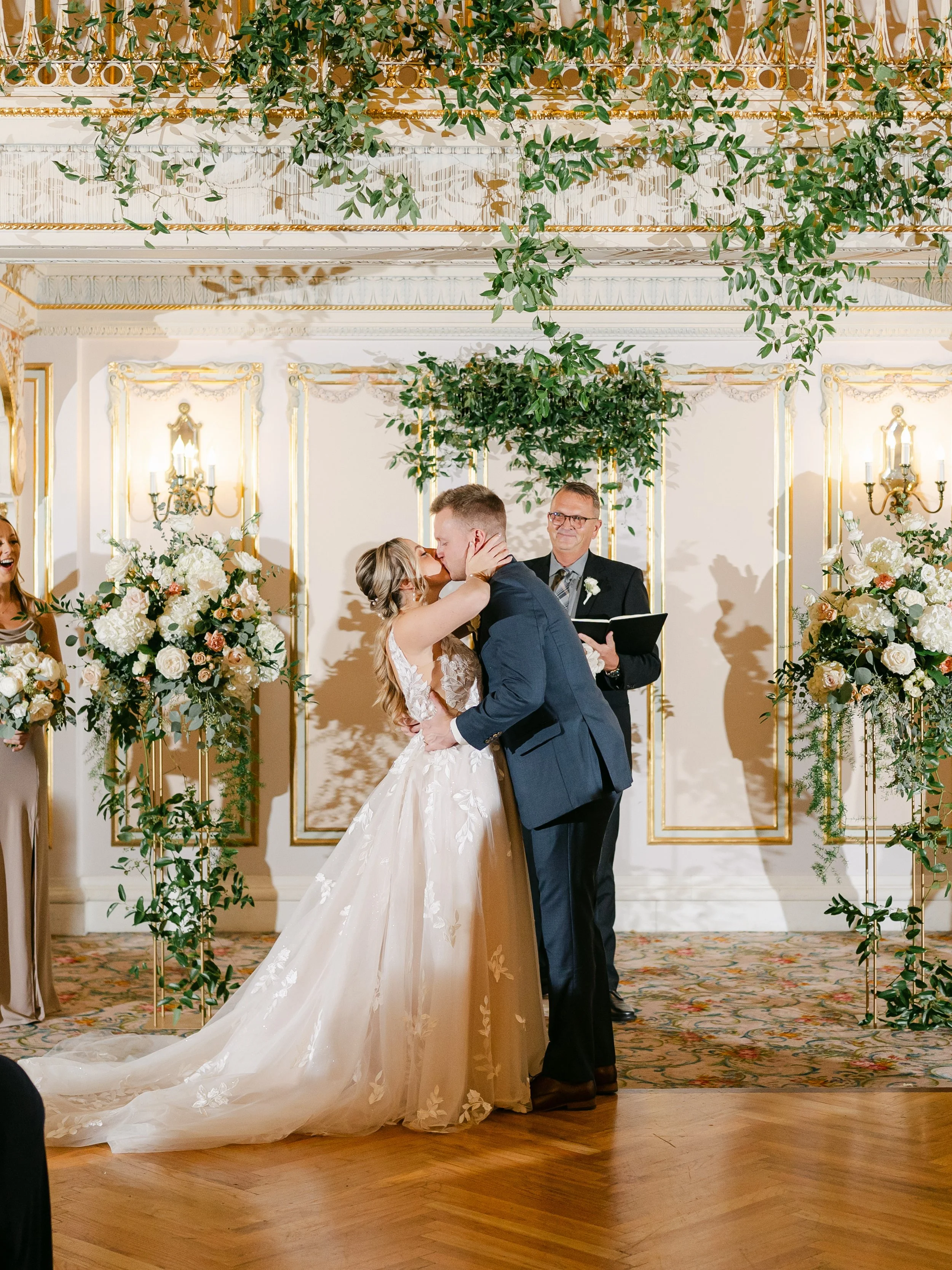 Bride and groom kiss during their wedding ceremony in an ornate ballroom decorated with greenery and white flowers.