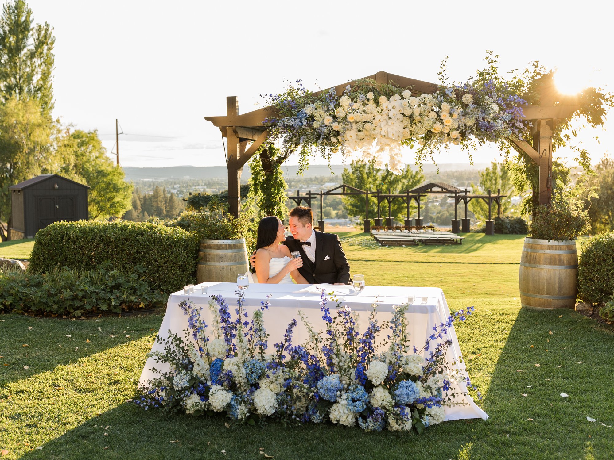 Bride and groom dining at a flower-adorned wedding table beneath a floral pergola at an outdoor venue