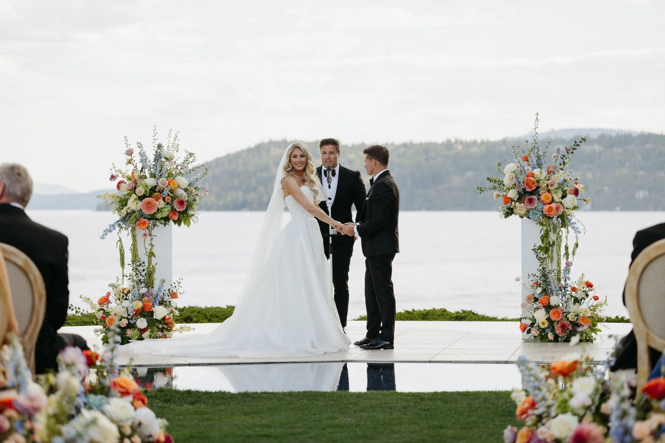 Bride and groom exchange vows beside a lake at an outdoor wedding ceremony with floral arrangements