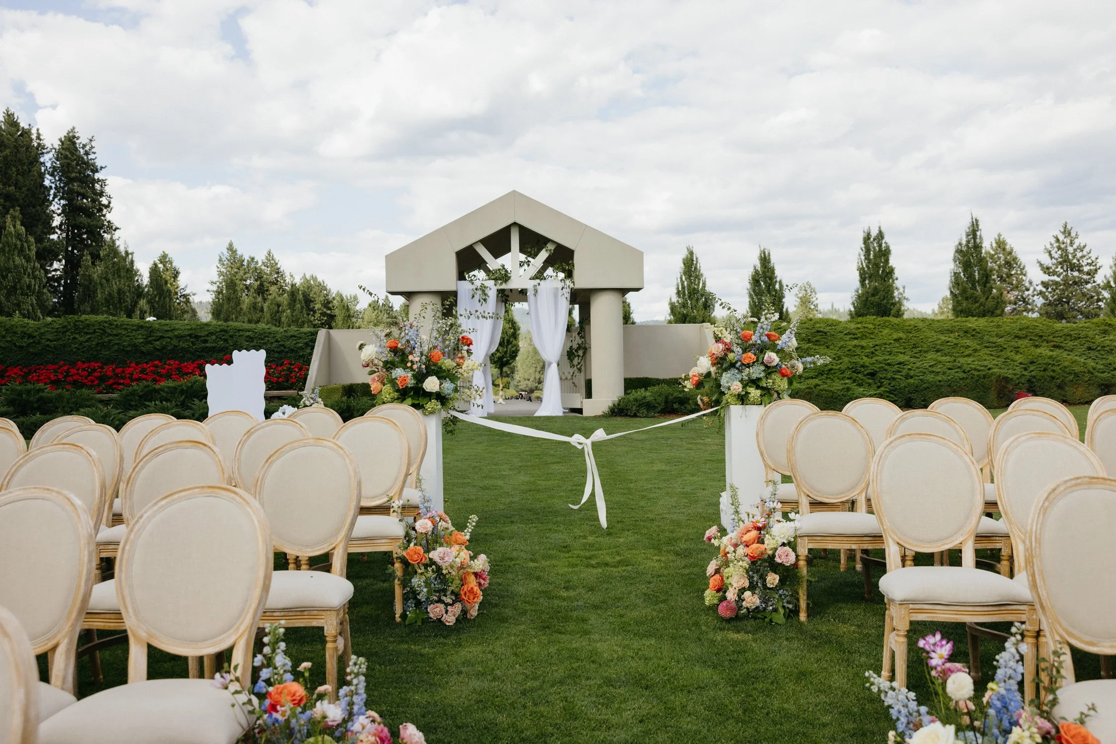 Outdoor wedding ceremony setup with floral arrangements, white drapery, aisle, and rows of cream chairs in a garden