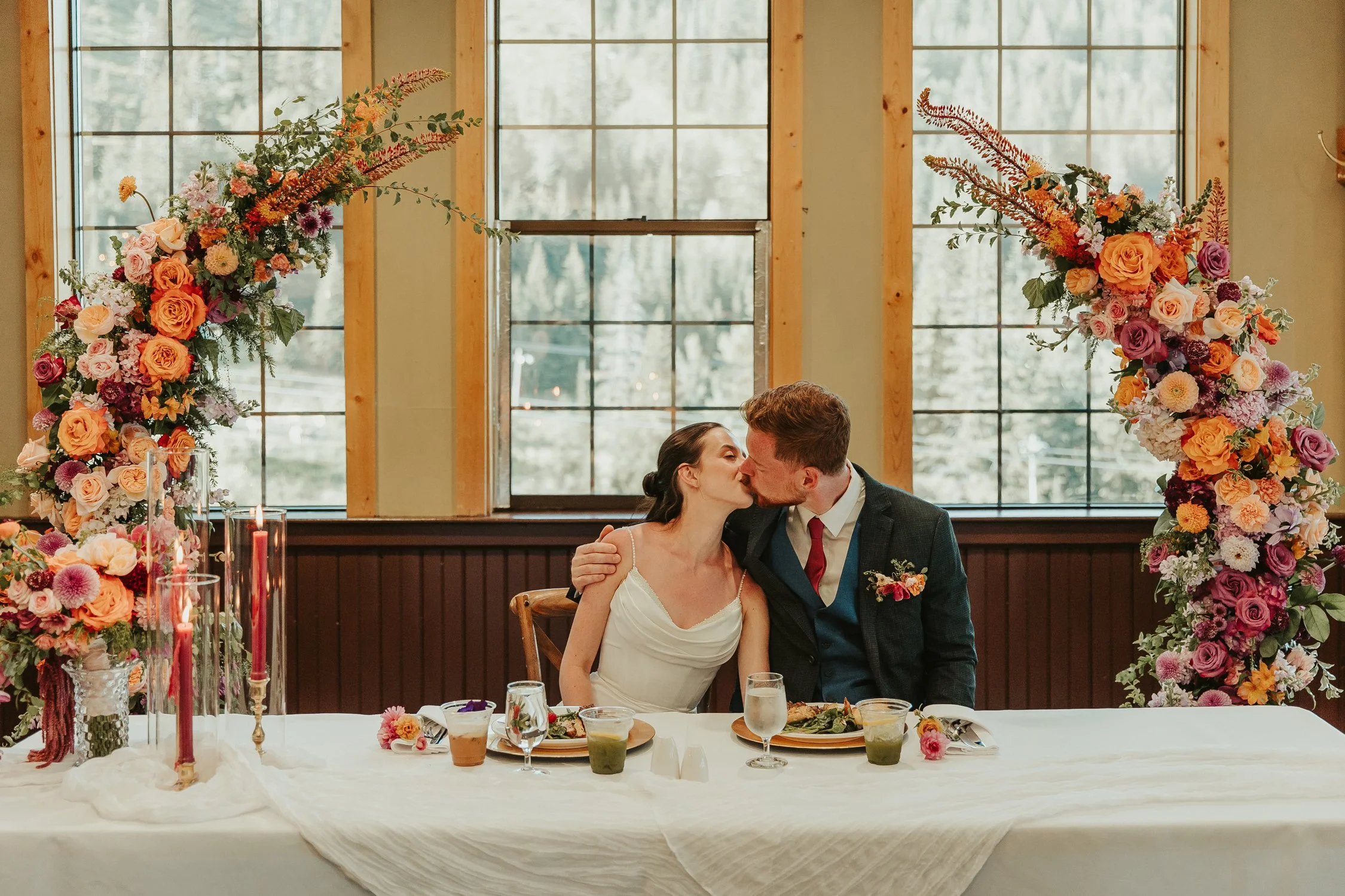 Newlywed couple kissing at a wedding reception dinner table surrounded by colorful floral arrangements and candles