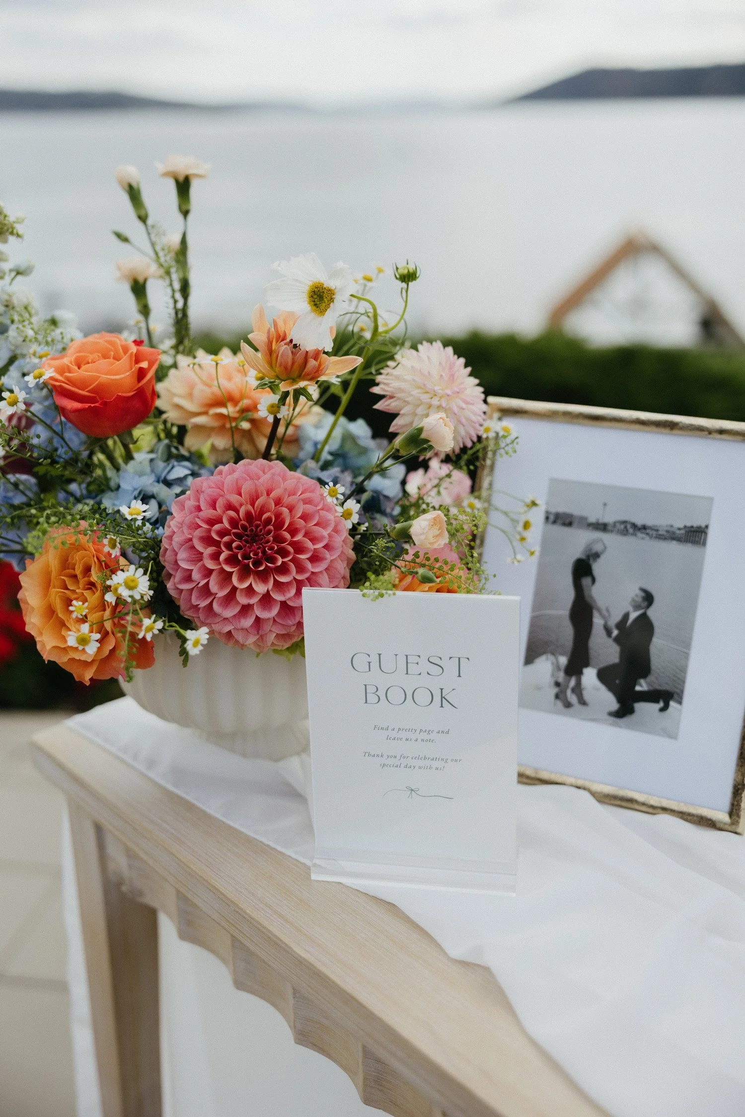 Wedding guest book beside a colorful floral arrangement and framed black-and-white wedding photo.