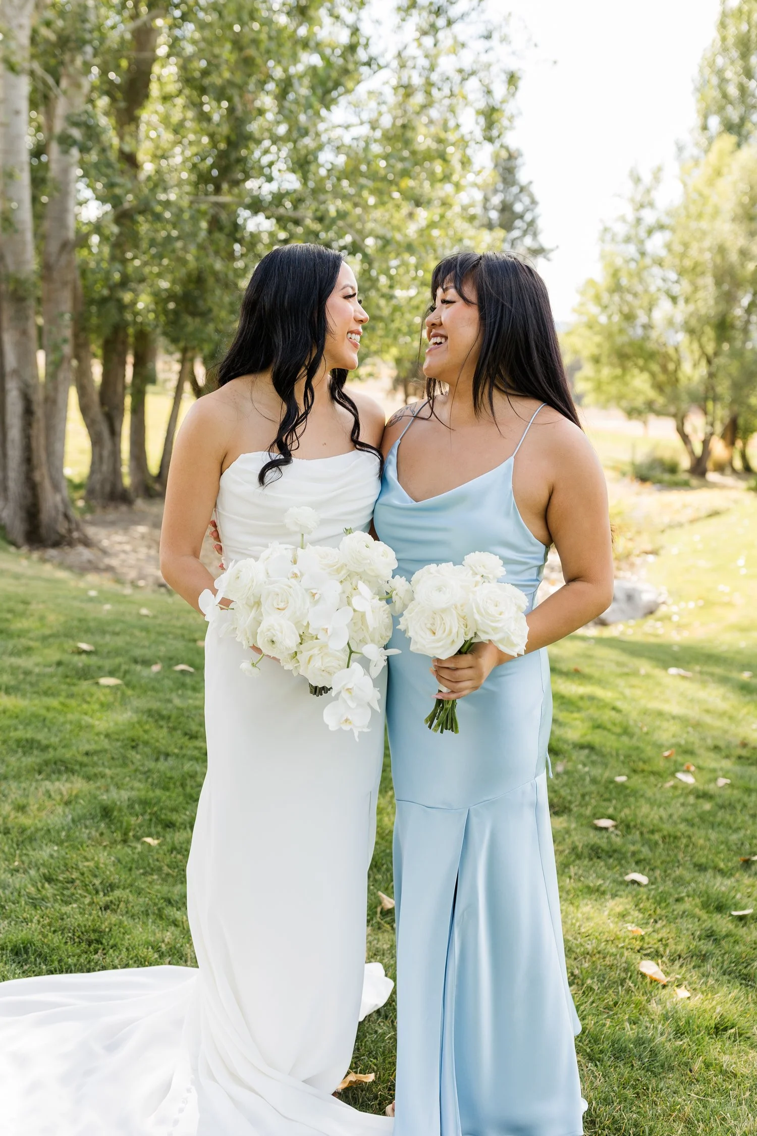 Two brides smiling together outdoors, holding white flower bouquets in a sunlit garden