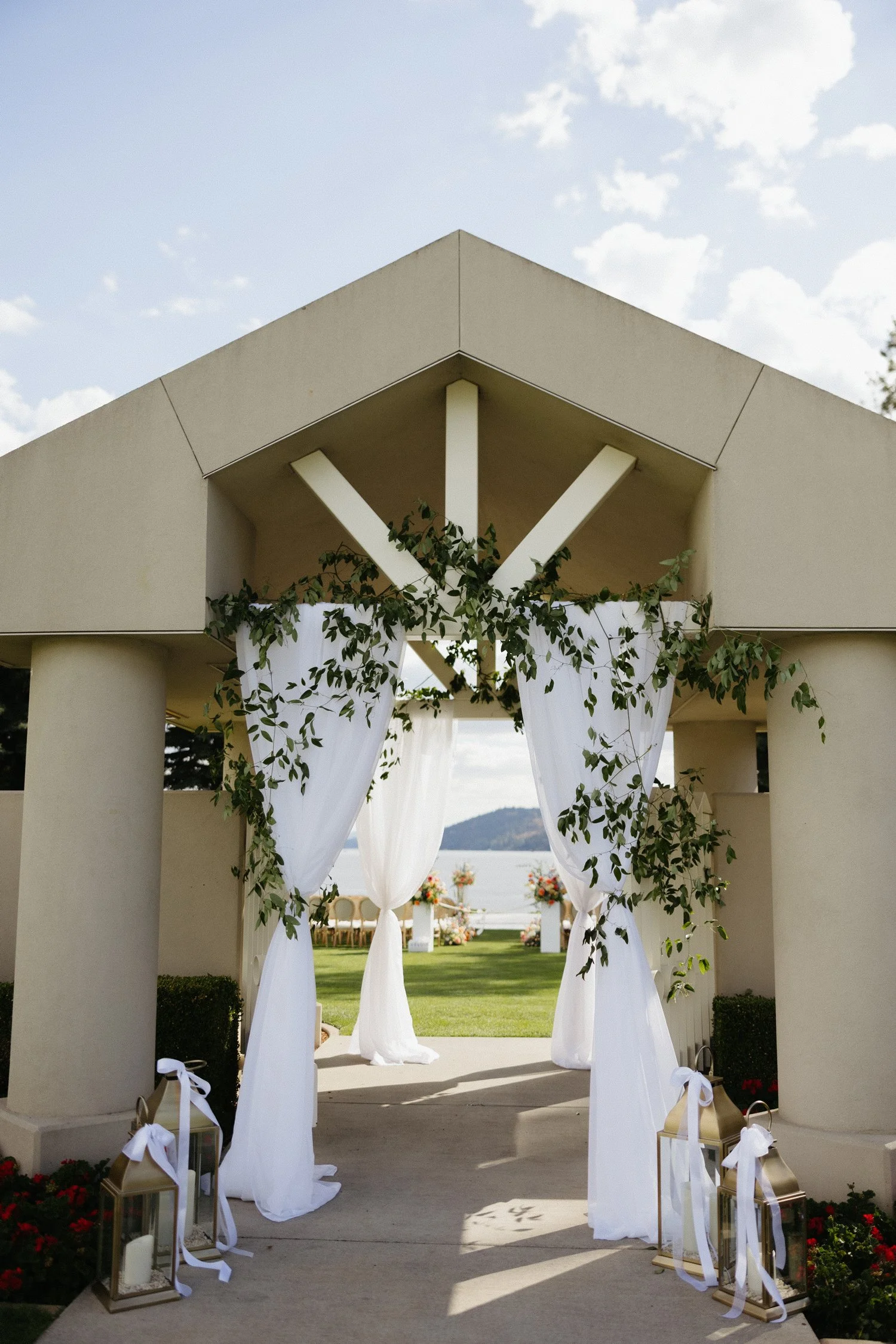 White-draped wedding ceremony entrance decorated with greenery, lanterns, and flowers overlooking a lake