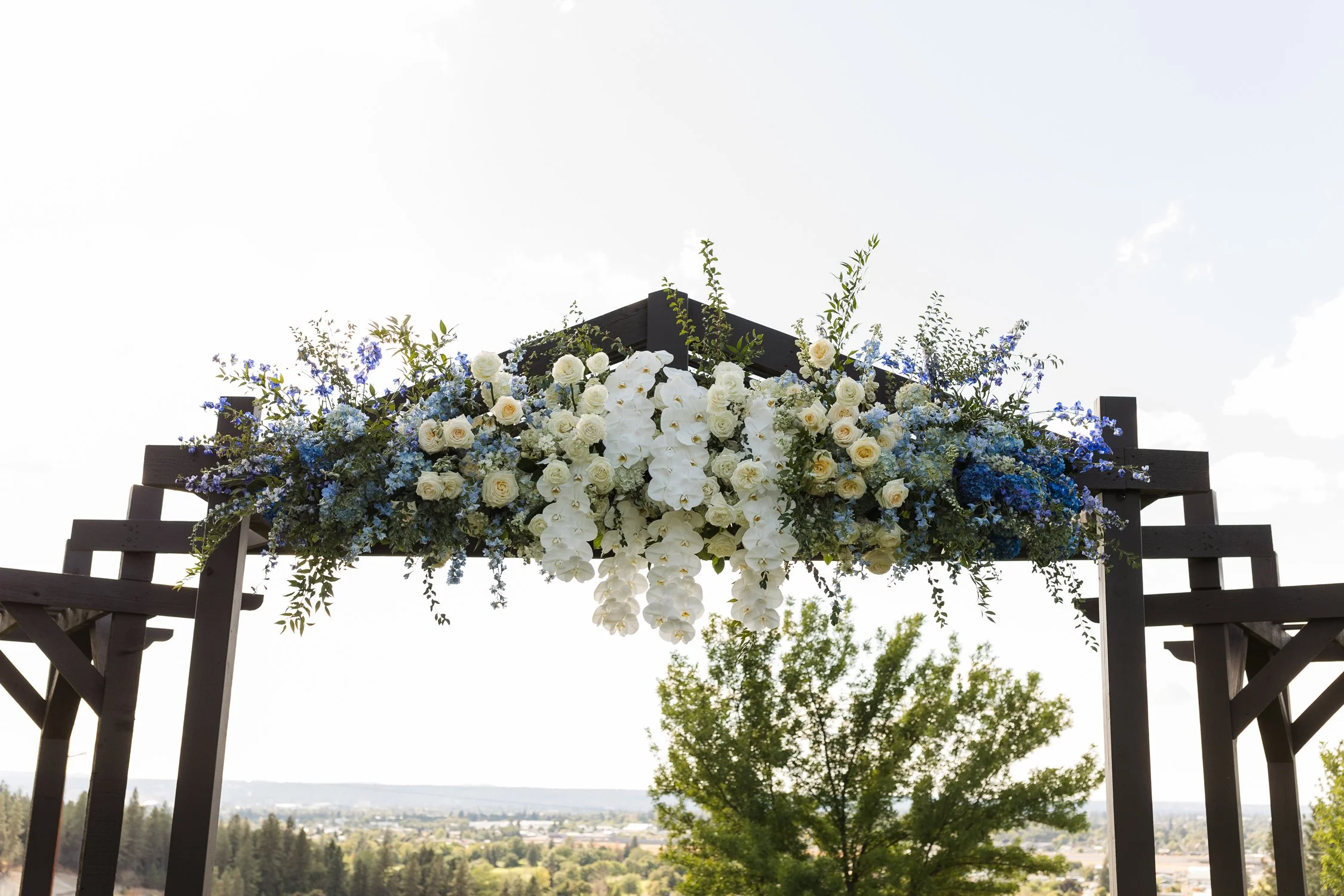 Wooden wedding arbor decorated with cascading white and blue flowers overlooking a scenic landscape