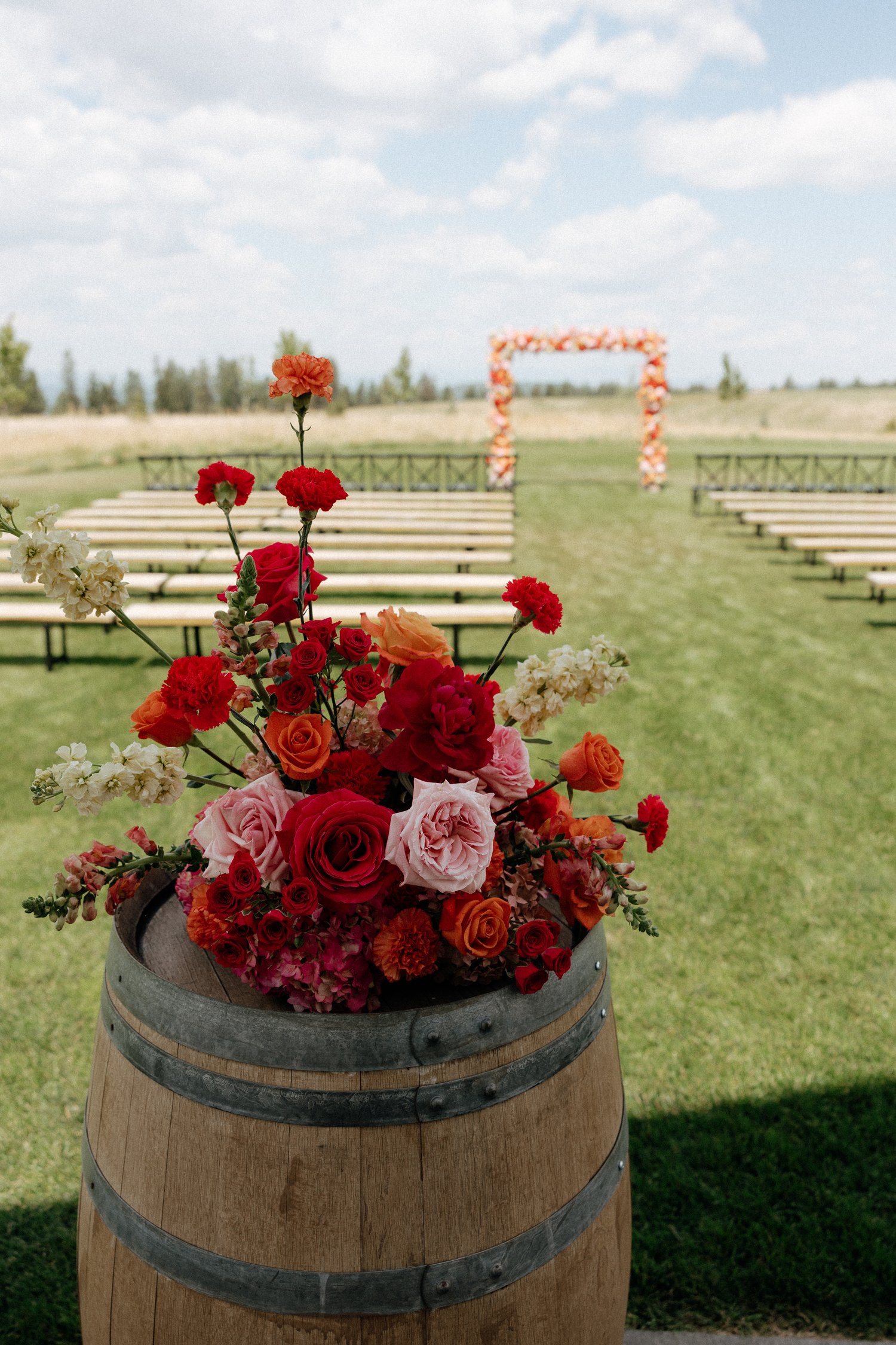 Colorful wedding flower arrangement in a wooden barrel overlooking outdoor ceremony seating and a floral arch