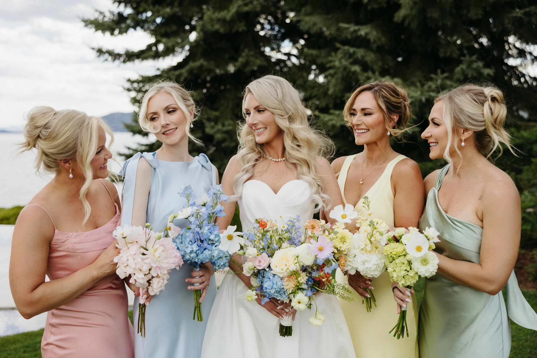 Bride and four bridesmaids holding colorful flower bouquets outdoors by a lake