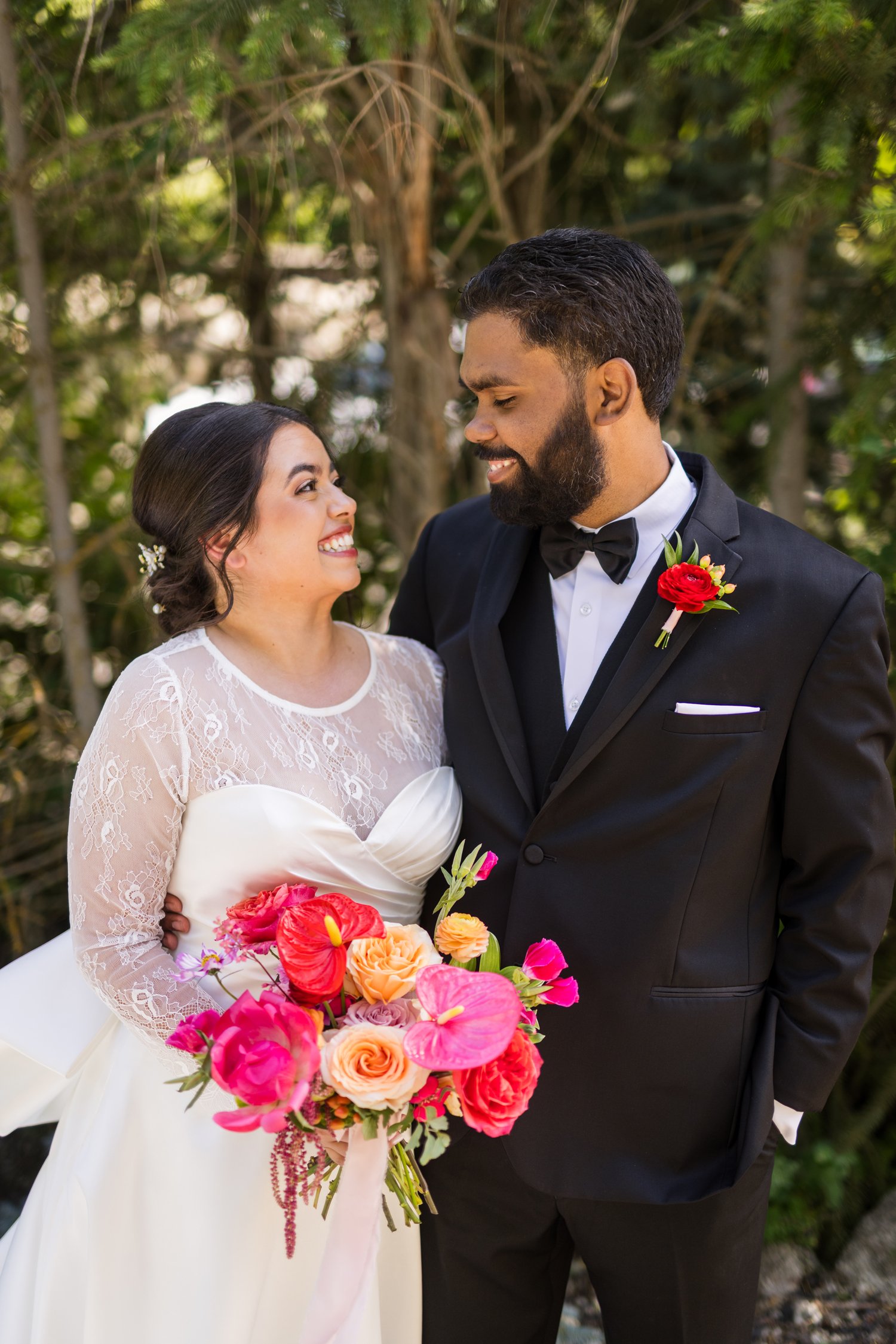 Newlywed couple smiling at each other outdoors, with the bride holding a colorful bouquet and the groom wearing a black tuxedo with a red boutonniere.