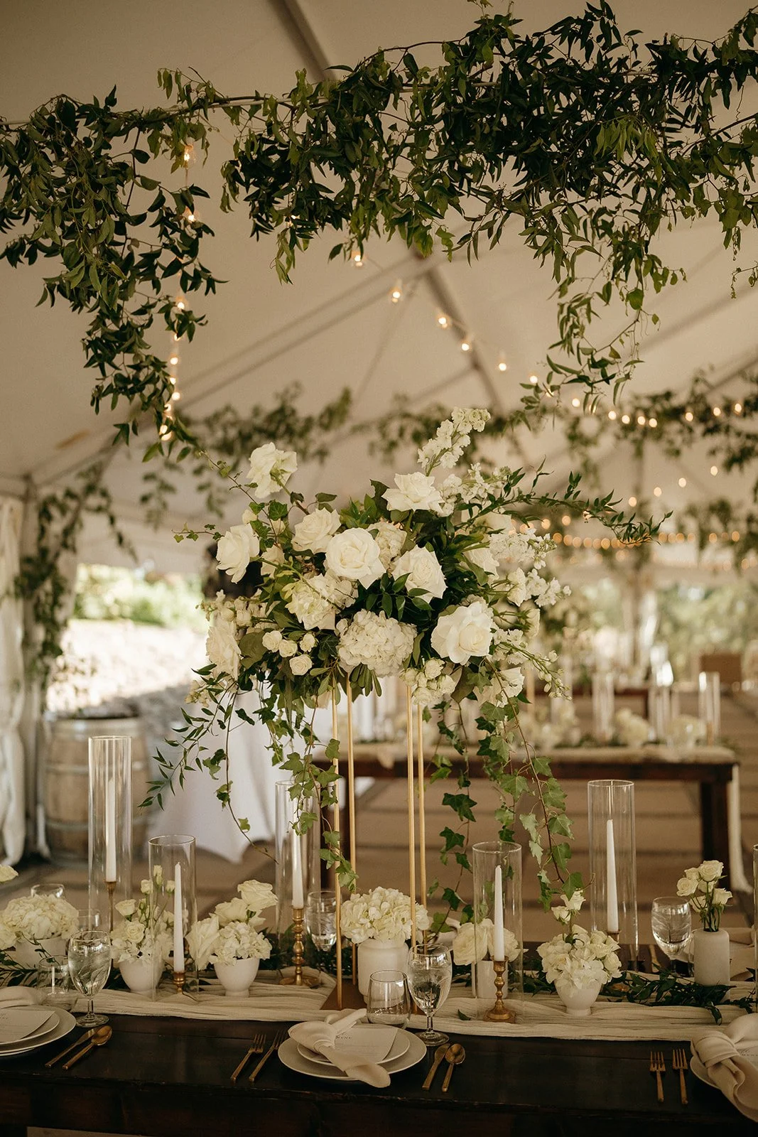Elegant wedding reception table with white roses, hydrangeas, greenery, candles, and warm string lights beneath a leafy canopy