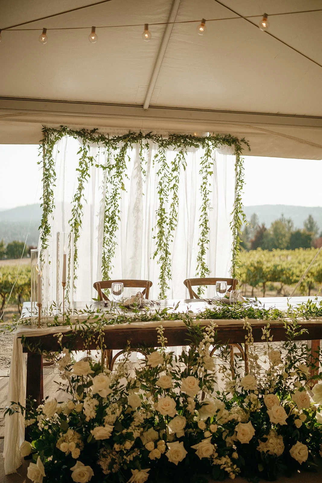 Elegant outdoor wedding reception table with white floral arrangement, greenery, sheer curtains, string lights, and vineyard view