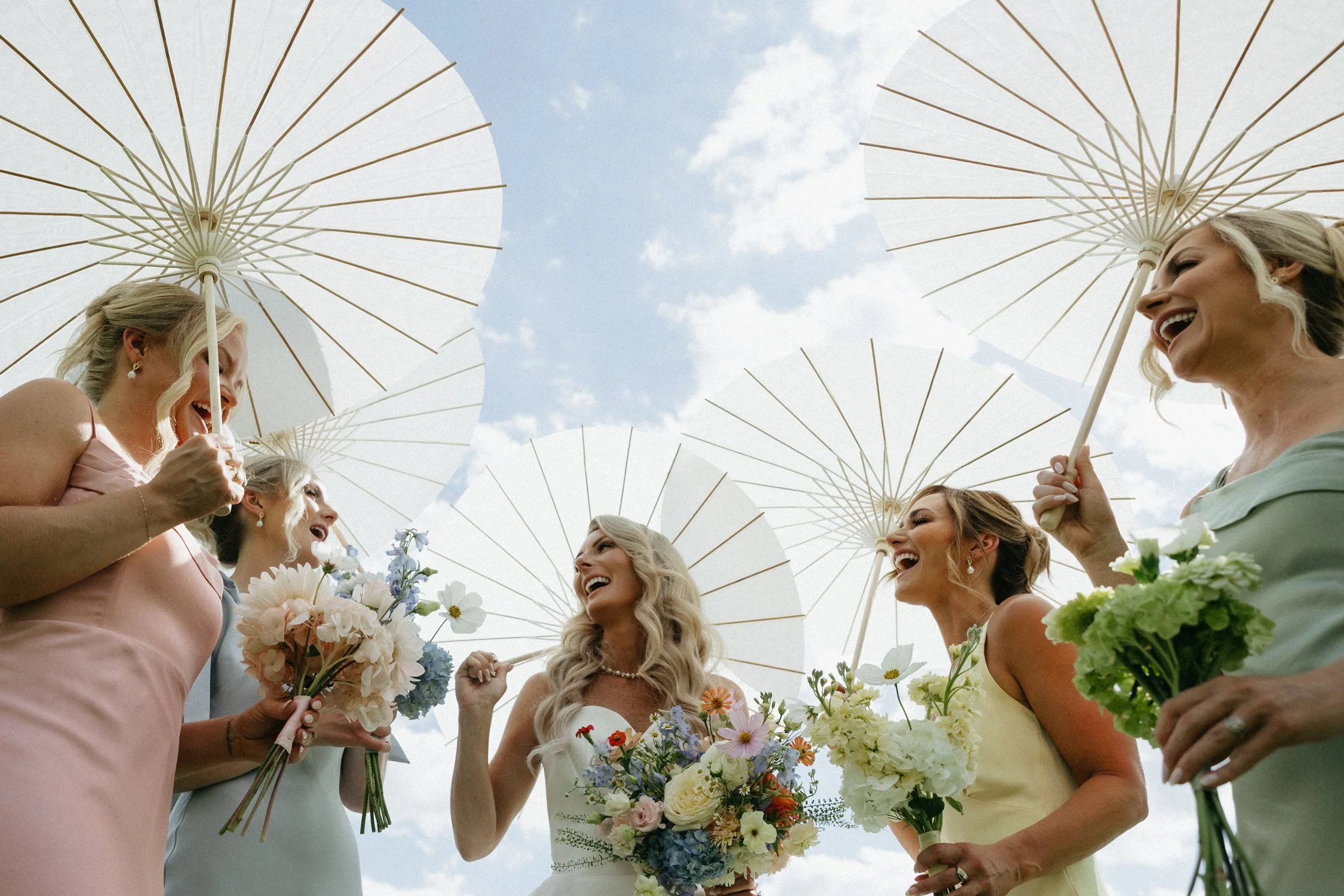Bride and bridesmaids laughing with bouquets and white parasols outdoors under a blue sky