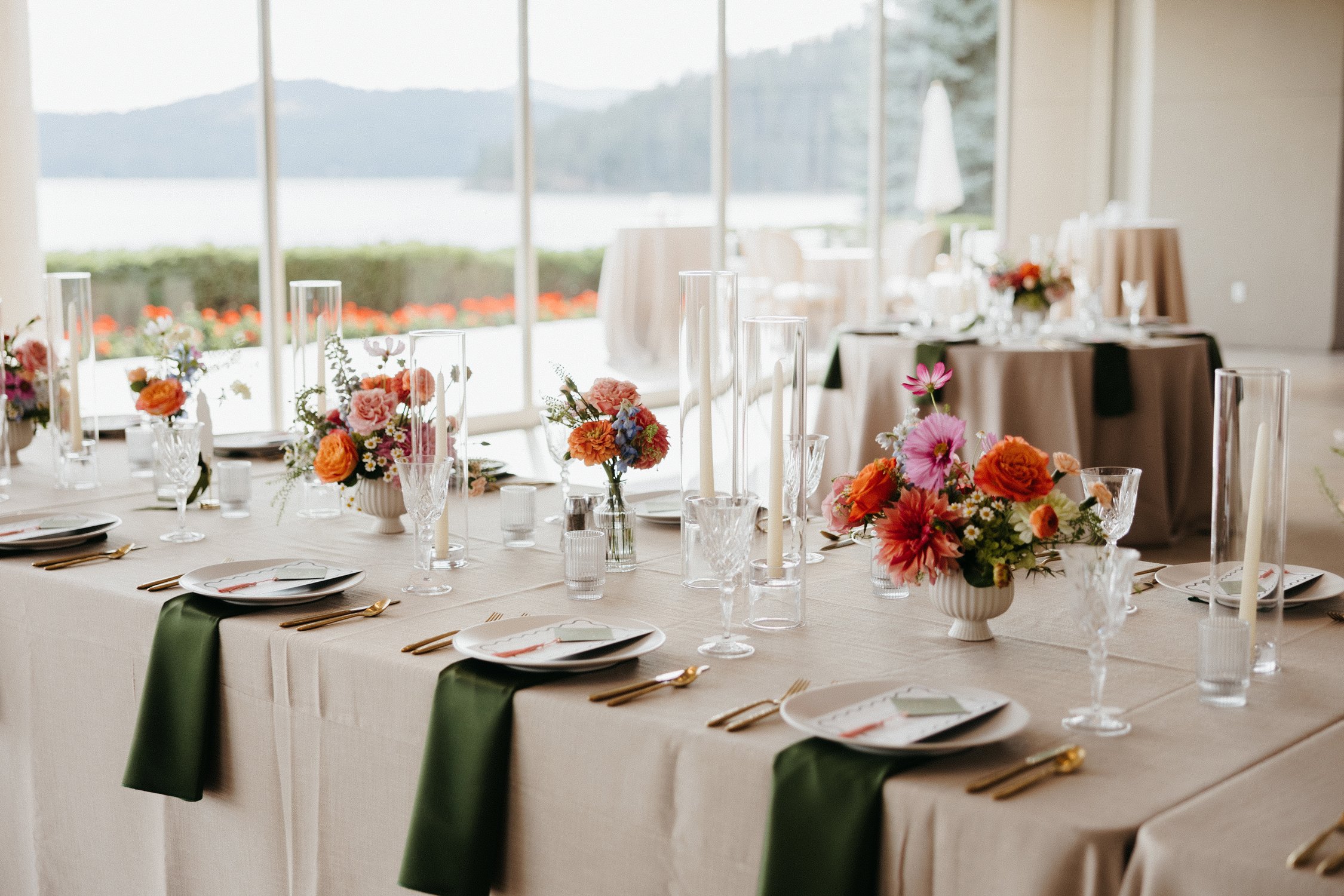 Elegant banquet tables with colorful floral centerpieces, candles, gold flatware, and green napkins overlooking a lake