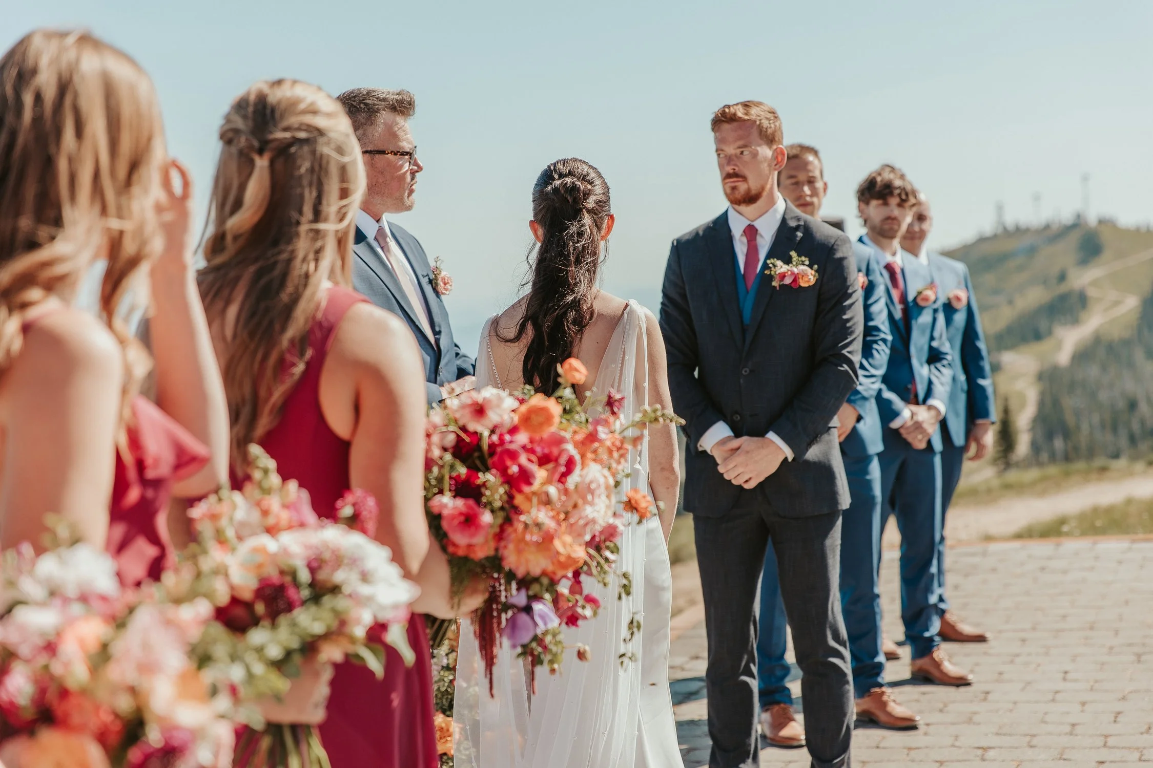 Outdoor wedding ceremony with bride, groom, bridesmaids, and groomsmen holding colorful bouquets against a mountain backdrop