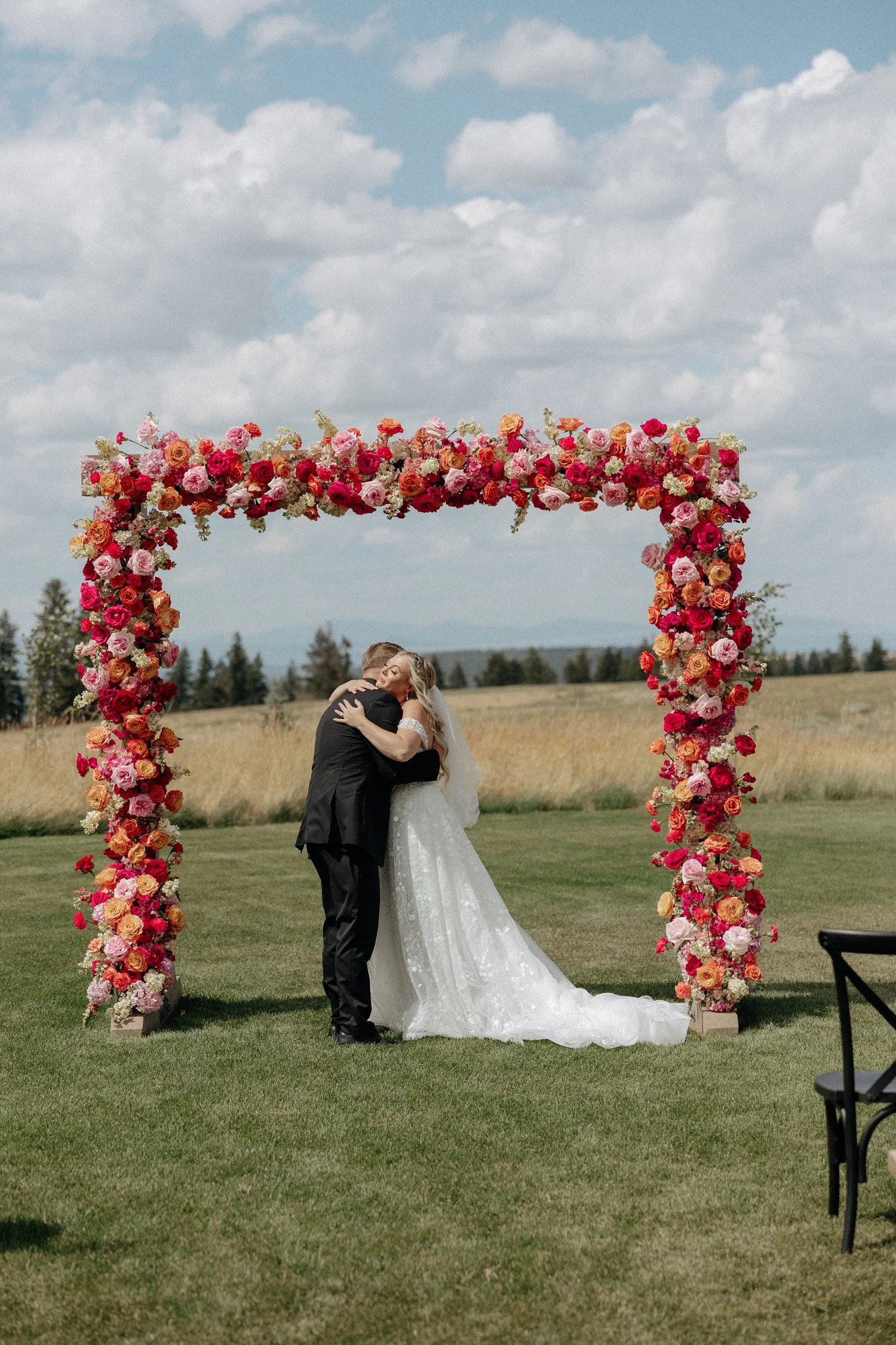 Bride and groom embracing beneath a colorful floral wedding arch in a sunlit field