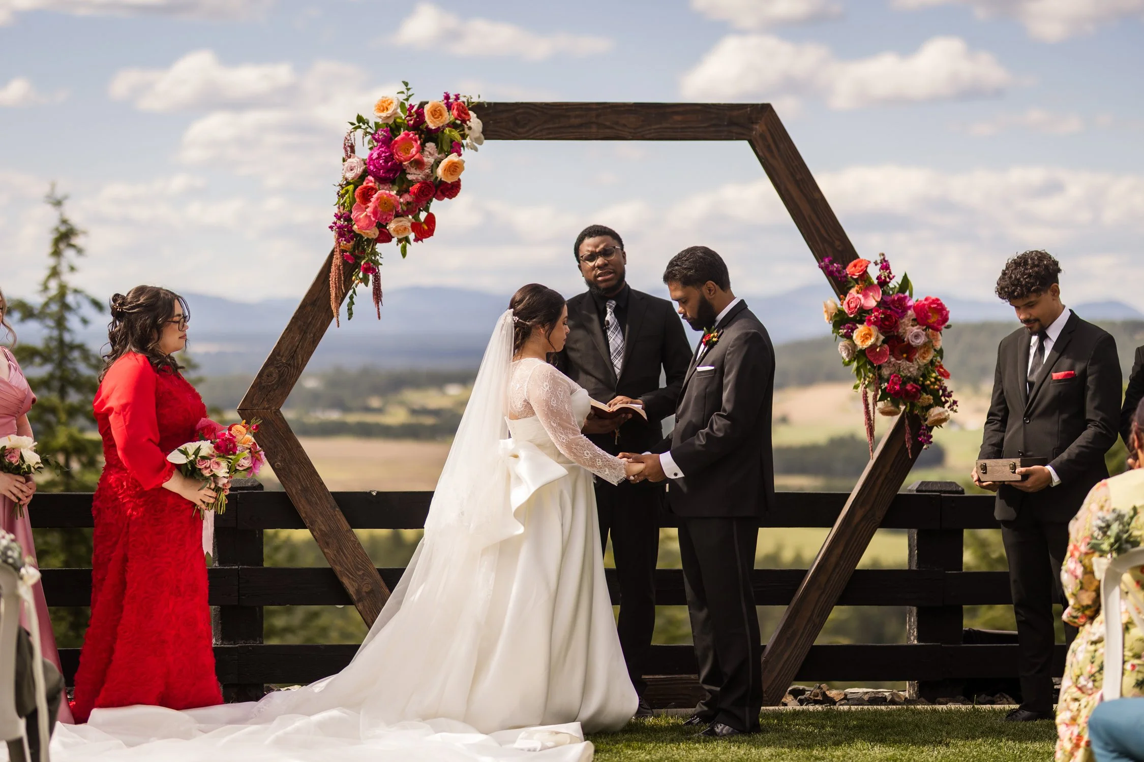 Outdoor wedding ceremony with bride and groom beneath a flower-adorned wooden arch, officiant, and wedding party.