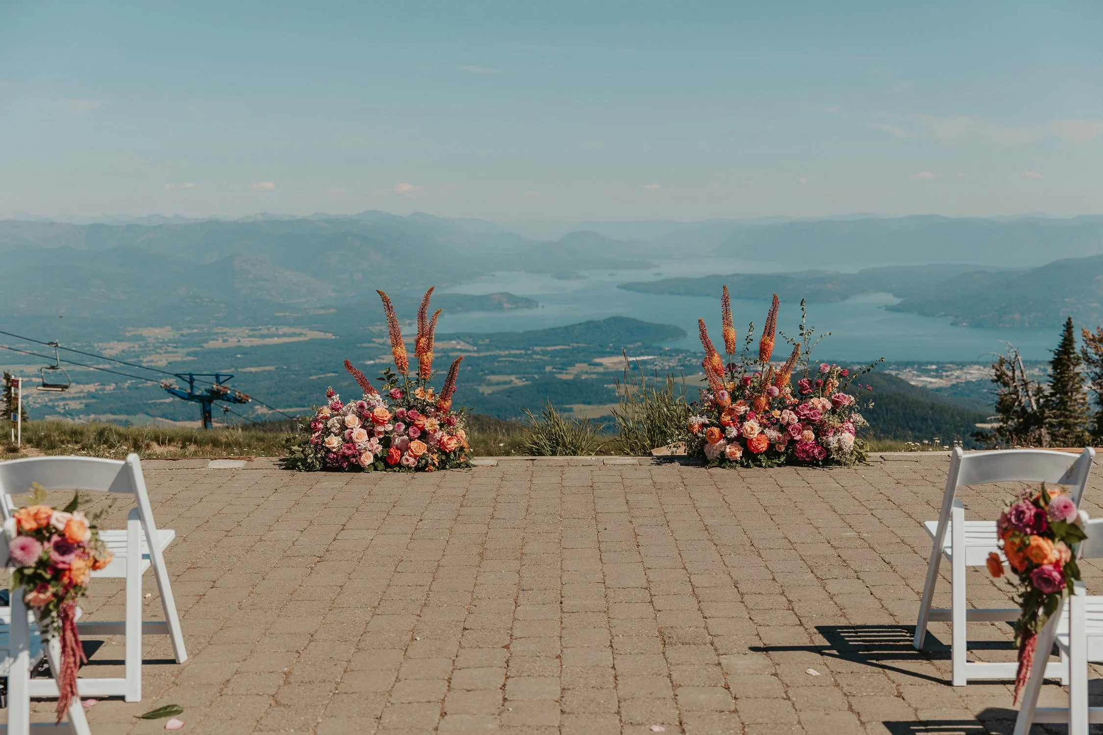Outdoor wedding ceremony setup with floral arrangements, white chairs, lake, mountains, and ski lift view