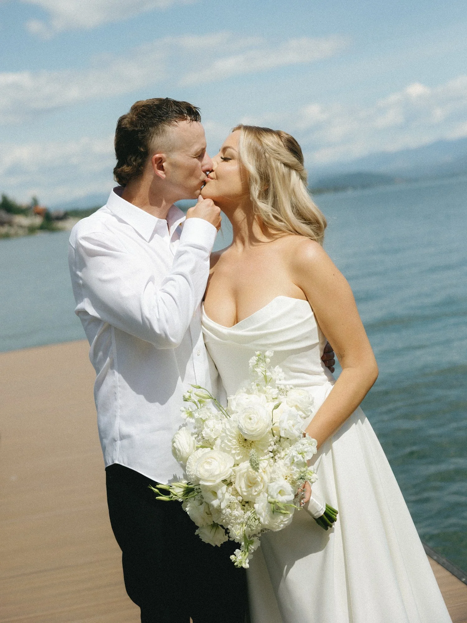 Newlywed couple kissing by the water, with the bride holding a white flower bouquet of white roses, ranunculus, stock flowers, hydrangea, and dahlias.
