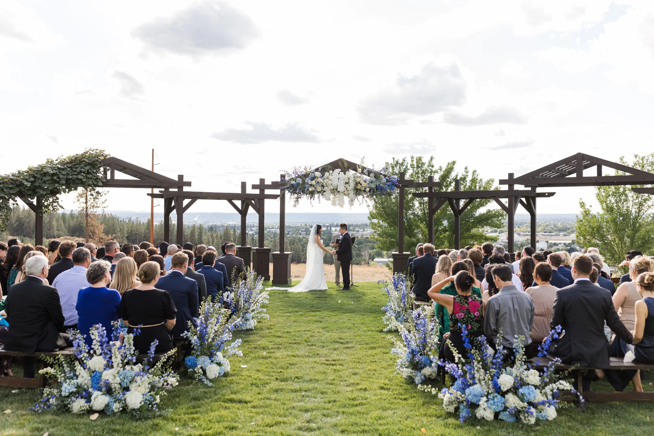 Outdoor wedding ceremony with bride and groom beneath a flower-adorned wooden arbor, surrounded by seated guests and mountain views