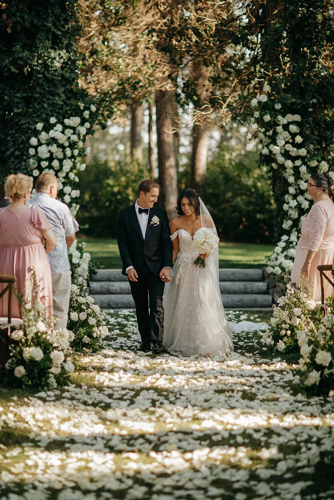 Bride and groom walking beneath a white flower-covered wedding arch in a forest garden