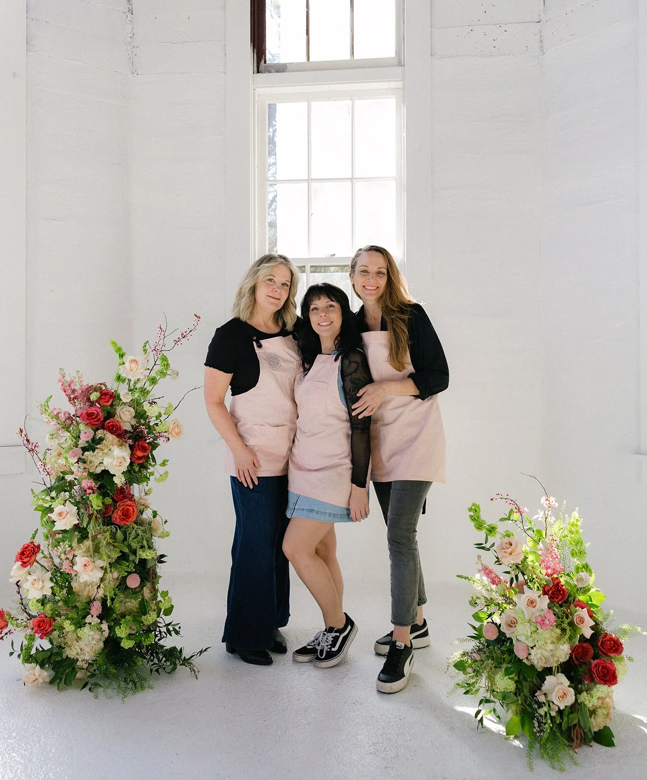 Three women in pink aprons pose between colorful floral arrangements in a bright white studio.