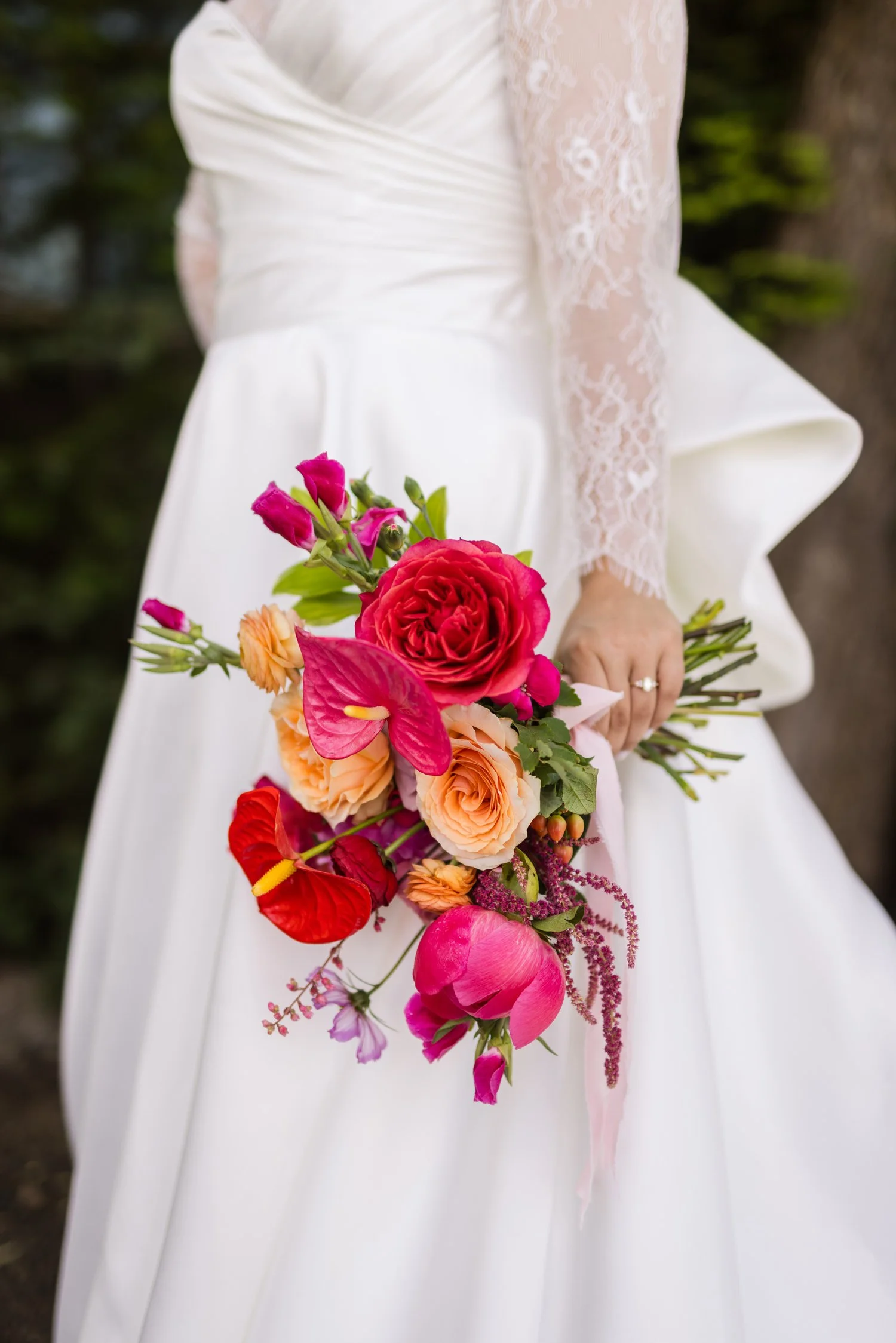 Bride in a white lace-sleeved wedding dress holding a colorful bouquet of roses, ranunculus, anthuriums, and peonies