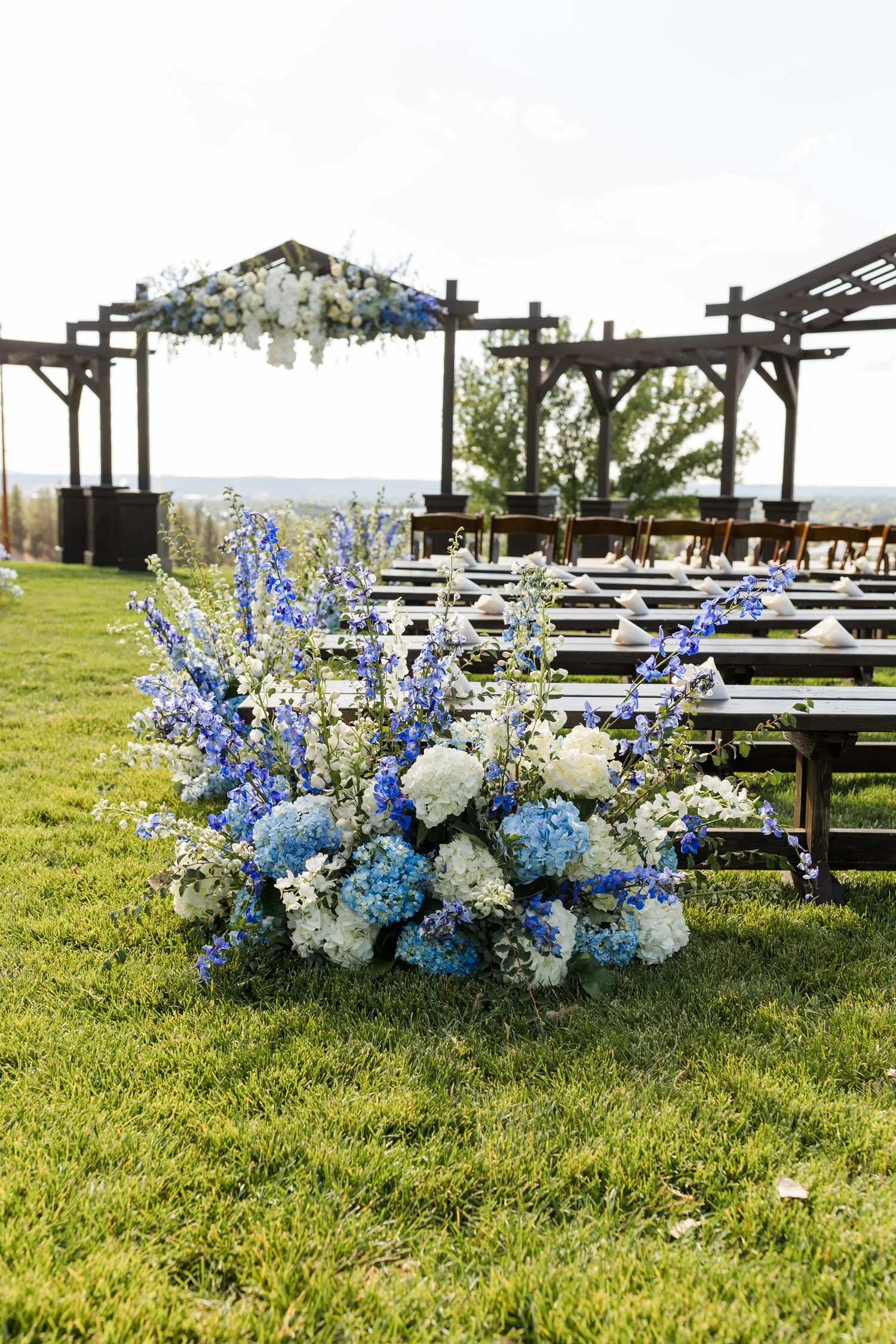 Outdoor wedding reception with blue and white floral arrangements, decorated picnic tables, and a wooden pergola overlooking the landscape