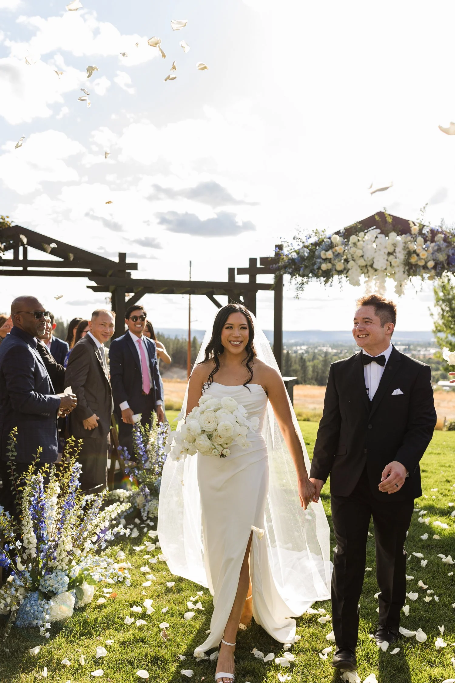 Bride and groom walking through an outdoor wedding ceremony as guests toss flower petals.