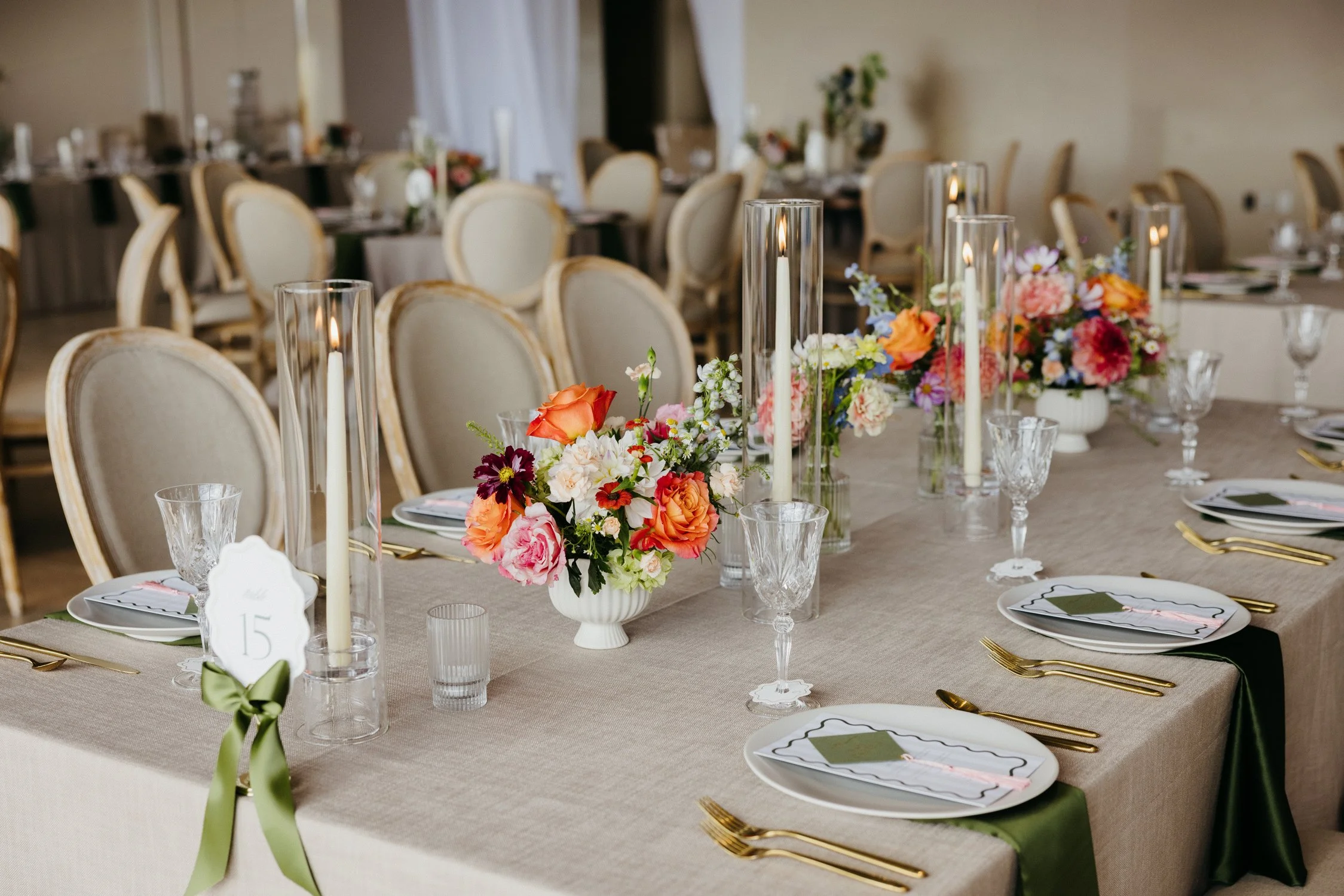 Elegant banquet table set with colorful floral centerpiece, taper candles, crystal glassware, gold flatware, and numbered place settings