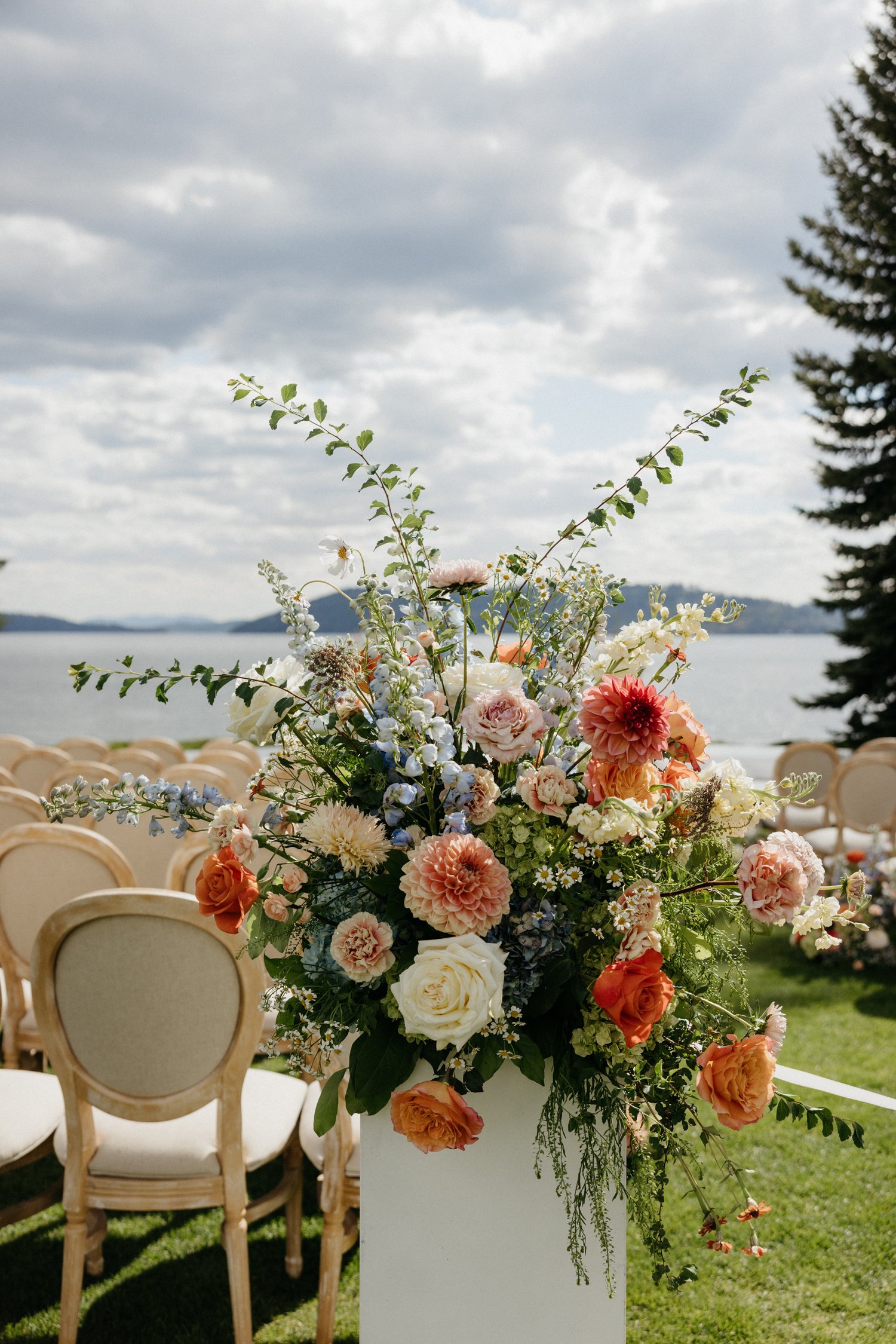 Colorful wedding flower arrangement on a pedestal overlooking a lakeside ceremony with wooden chairs and mountains.