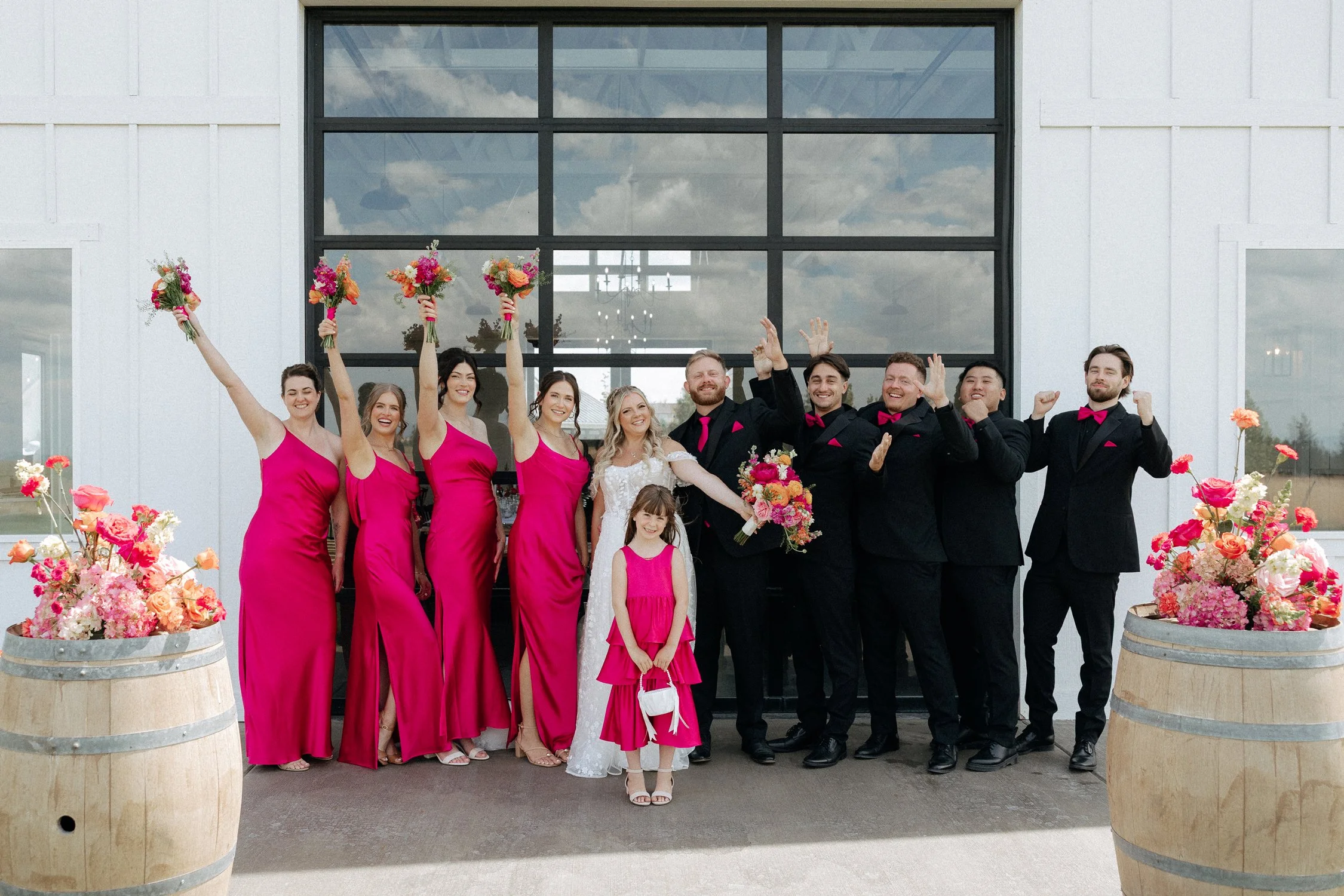 Wedding party posing outside with bridesmaids in pink dresses, groomsmen in black suits, and colorful flowers