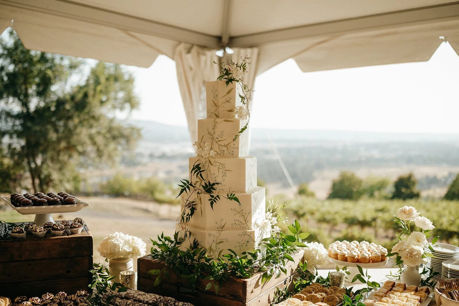 Elegant tiered wedding cake decorated with white flowers and greenery at an outdoor dessert table with cupcakes and pastries.