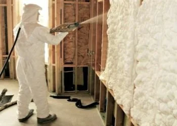 Person in protective gear applying spray foam insulation to a wall in a building under construction.